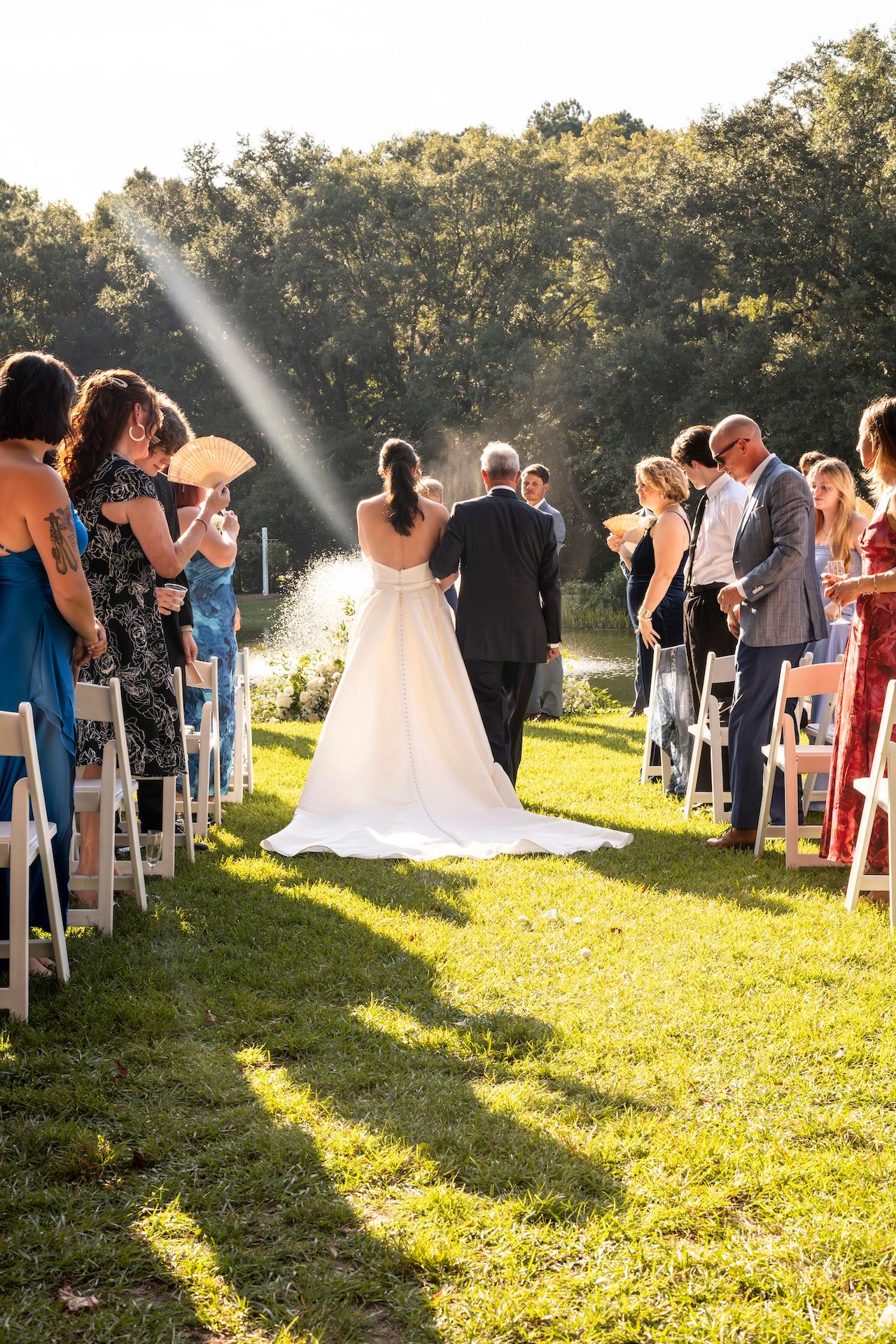 An outdoor wedding ceremony on a sunny afternoon with guests seated in white chairs while a ray of sunlight beams down.