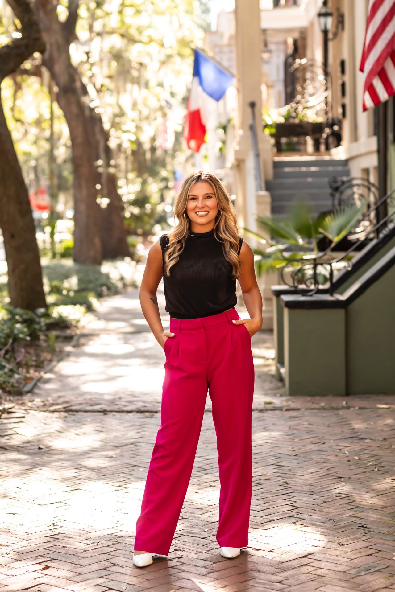 A woman in a black sleeveless top and bright red wide-leg pants stands on a brick sidewalk with American flags in background.