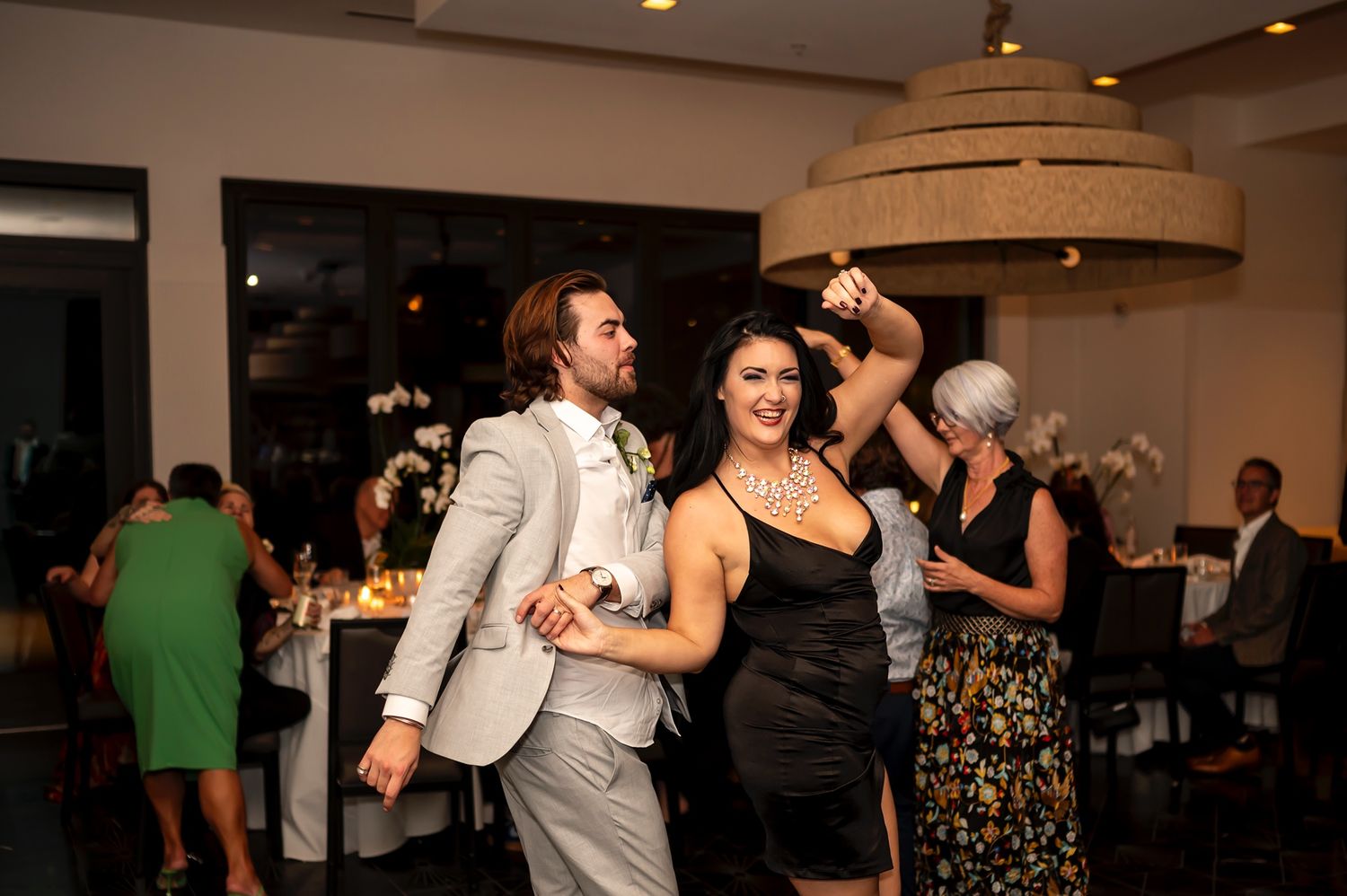 People dancing together at an indoor reception with low lighting and a bright chandelier in the background.