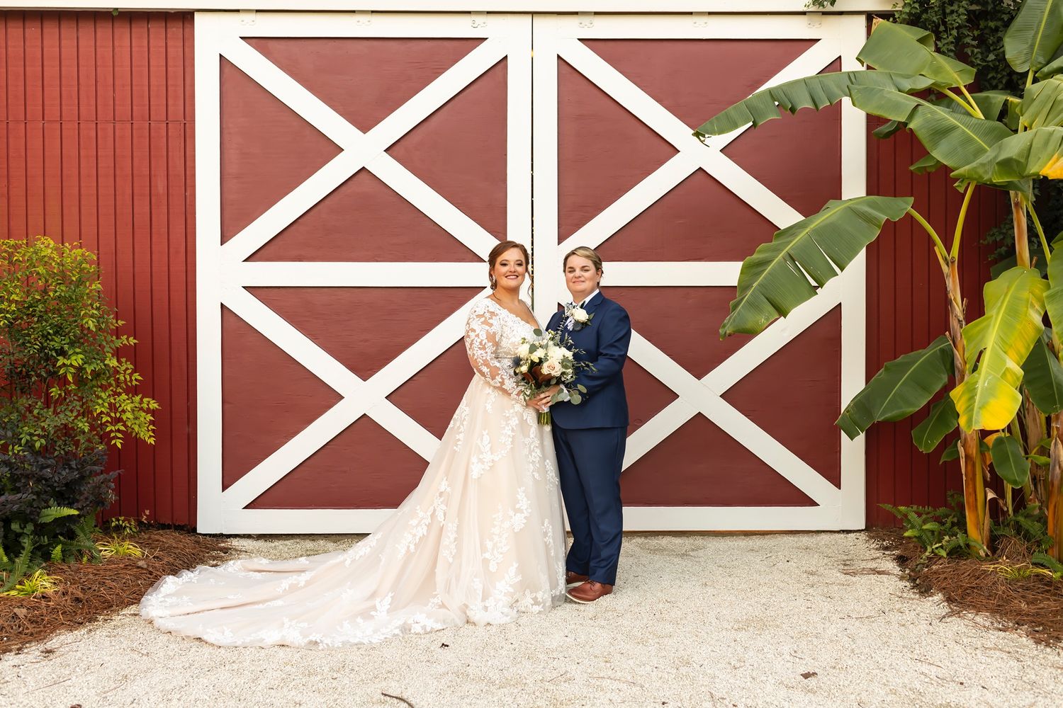 A couple stands together in front of large red barn doors with white crossbeams at a rustic farm wedding venue.