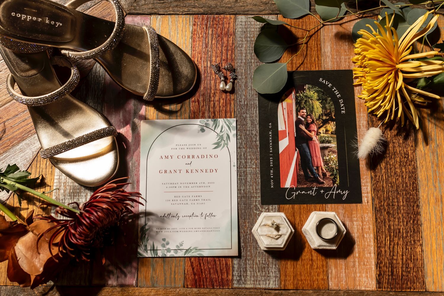 Wedding stationery and details arranged on a wooden table with sandals, flowers, and hexagonal ring boxes.