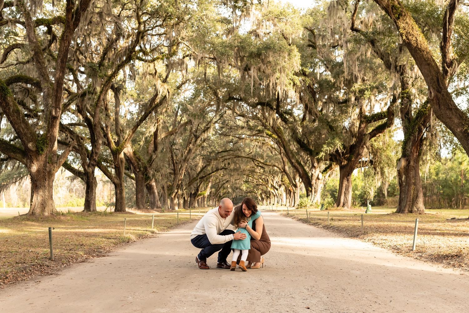 Two people and a dog share a tender moment on a path under a canopy of moss-draped oak trees.