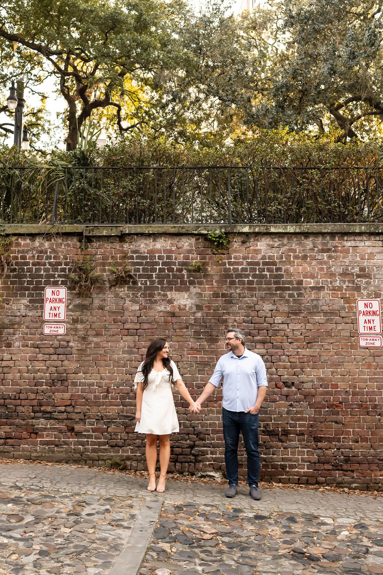 A couple holds hands against a rustic brick wall with cobblestone ground and overhanging trees at sunset.