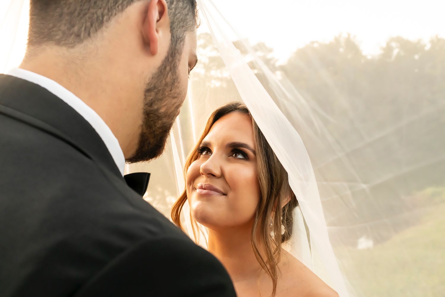 Smiling bride in lace veil gazes lovingly at groom during outdoor wedding ceremony at sunset.