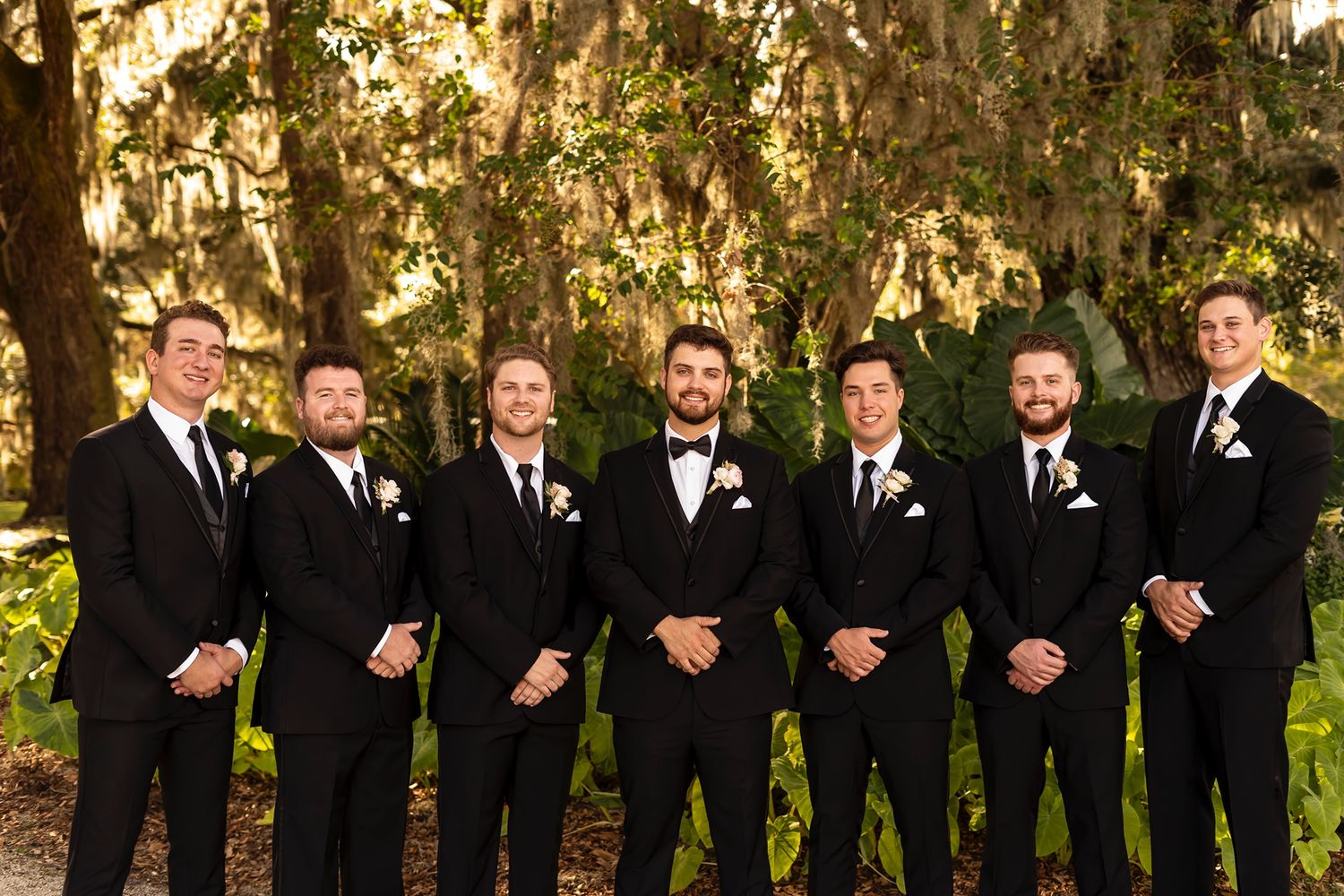 Groomsmen in classic black tuxedos and suits stand together outdoors under Spanish moss hanging from trees.