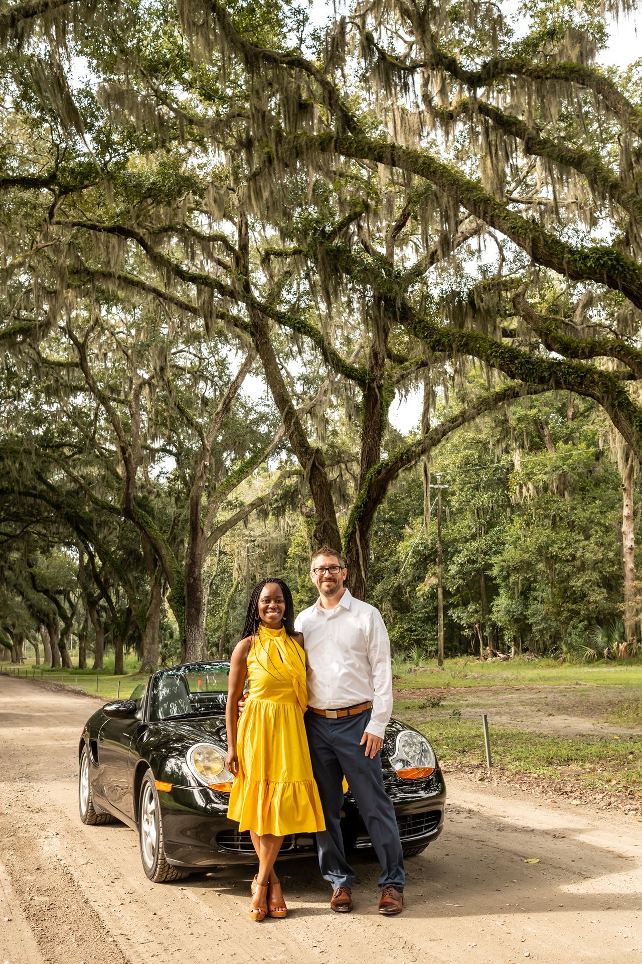 A couple poses in front of Spanish moss draped oak trees and a black car on a dirt road in an outdoor setting.