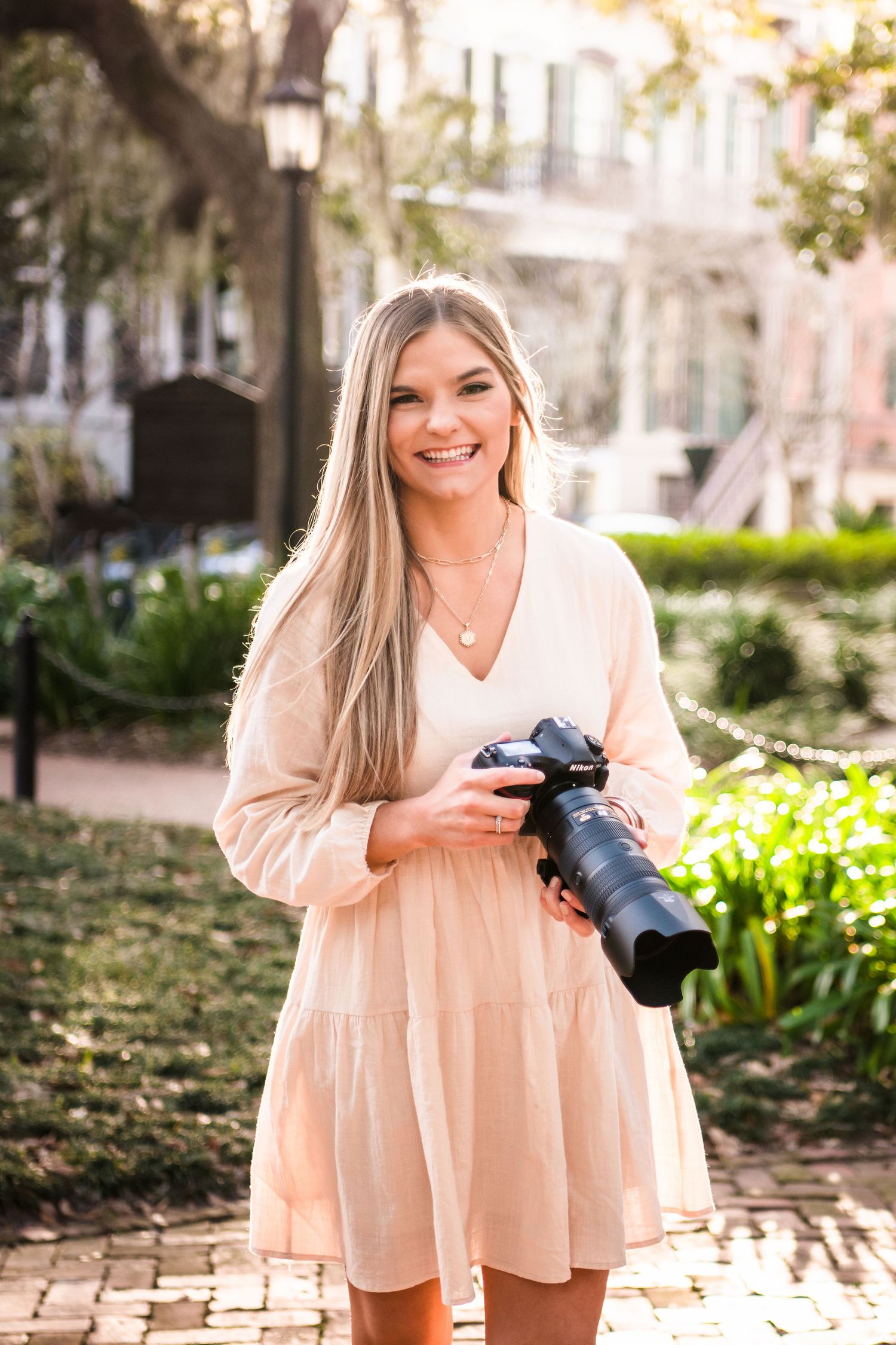 A photographer in a flowy peach dress smiles while holding a professional camera in a sunlit garden setting.