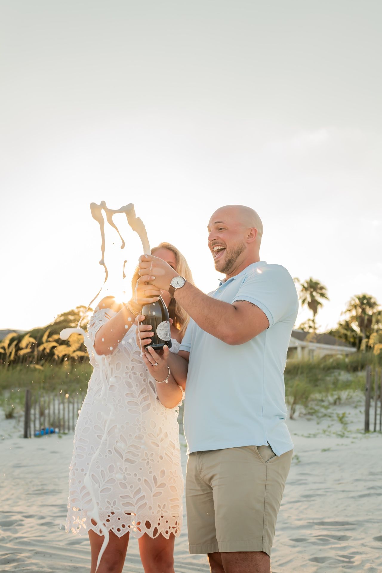 A joyful couple celebrates by popping champagne on a beach at sunset with palm trees in the background.