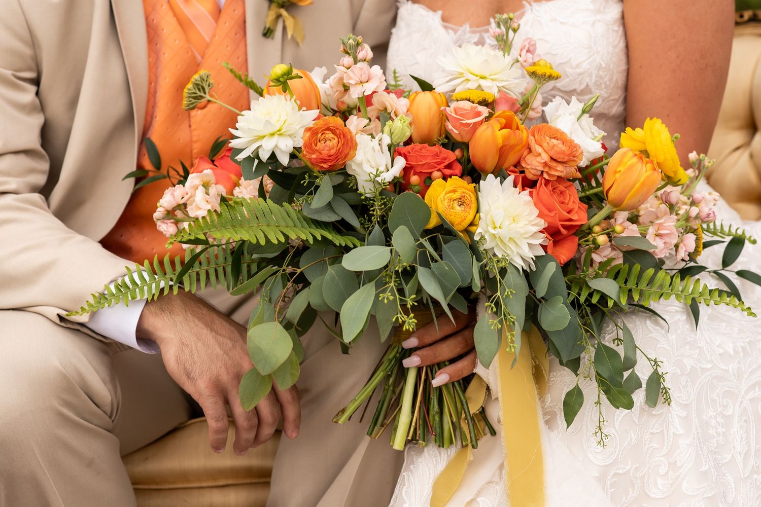 Vibrant wedding bouquet with orange, yellow and white flowers mixed with ferns and greenery held at a ceremony.