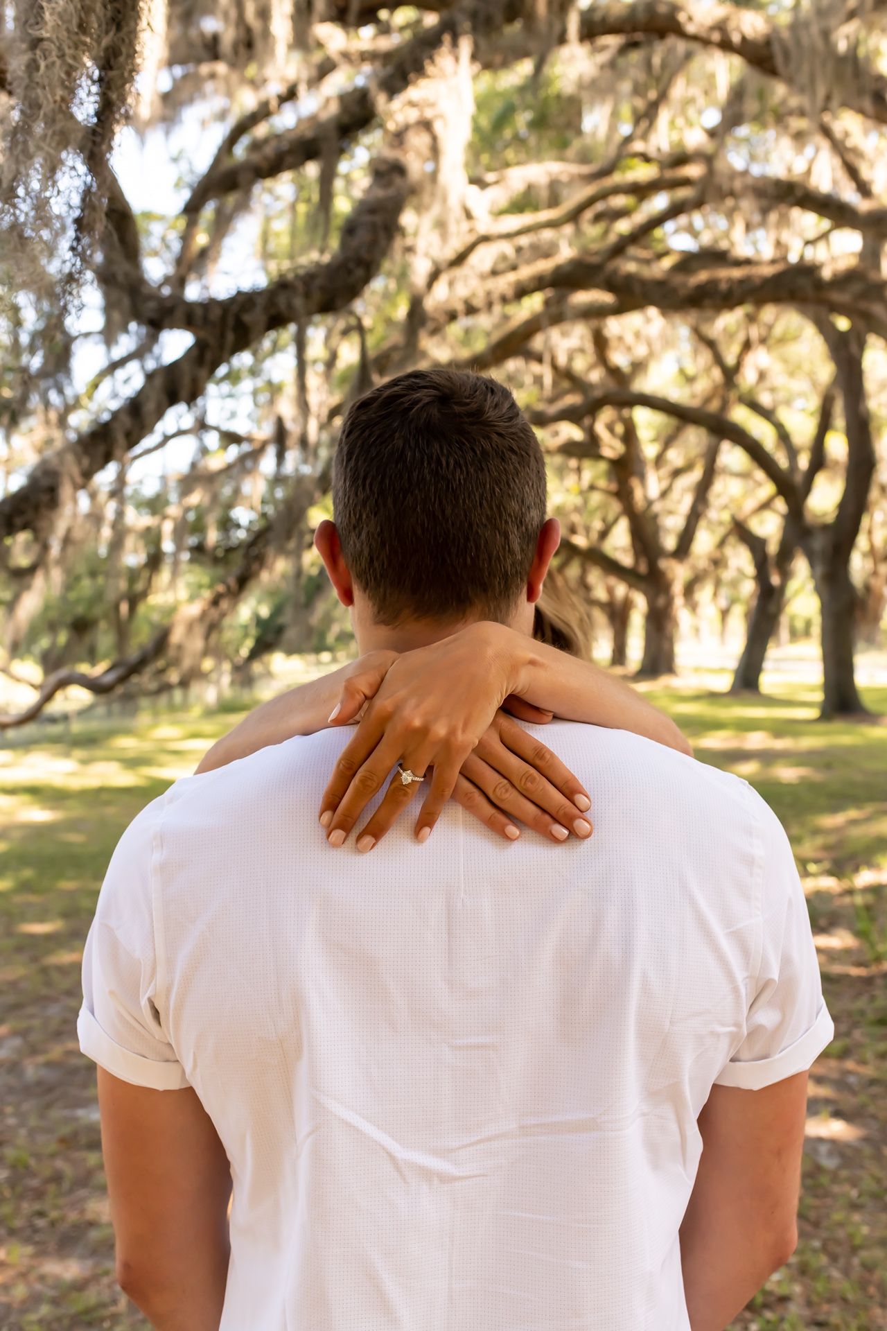 A person in a white t-shirt stands in front of moss-draped oak trees while hands rest on their shoulders from behind.