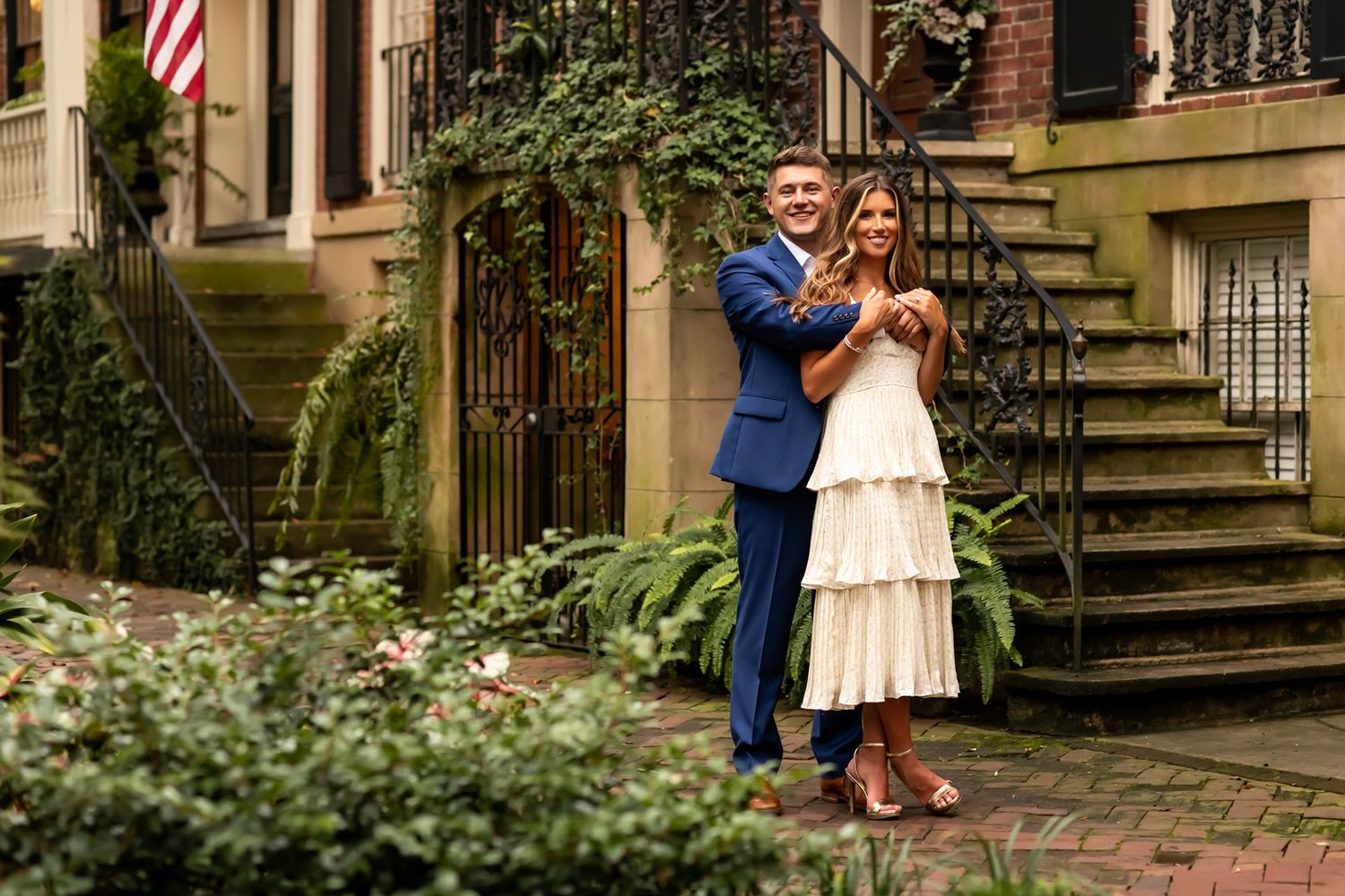 A couple in formal attire stands together on the steps of a brownstone building with ivy and garden landscaping.