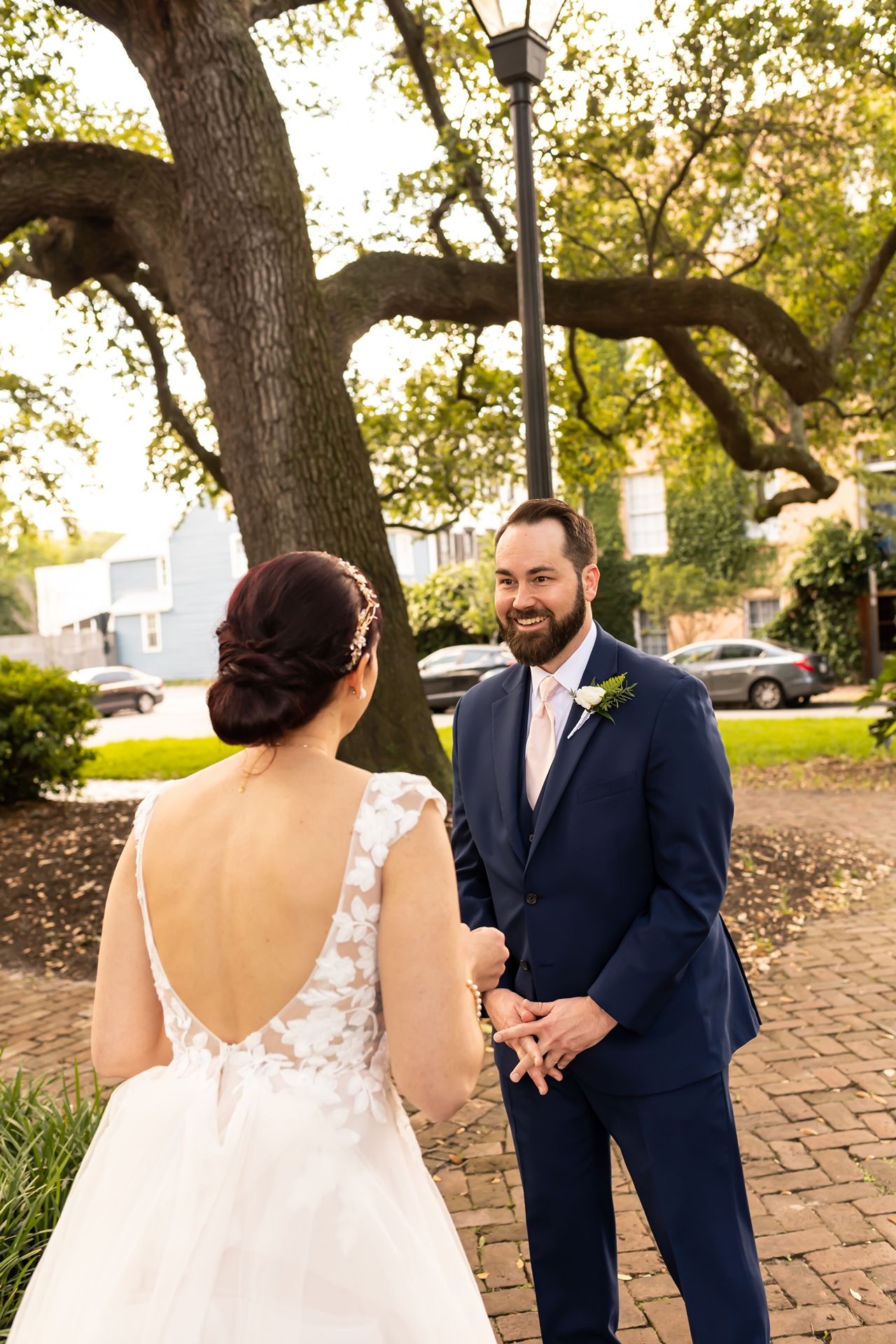Two people in wedding attire stand on a brick pathway under a large oak tree during golden hour sunset.