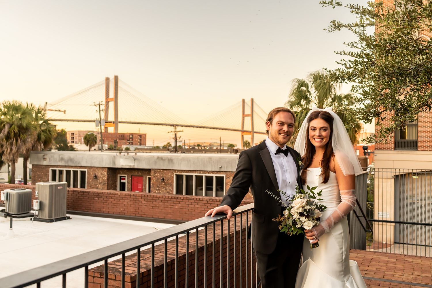 Elegant wedding couple poses on rooftop terrace at sunset with Savannah's Talmadge Bridge in background.
