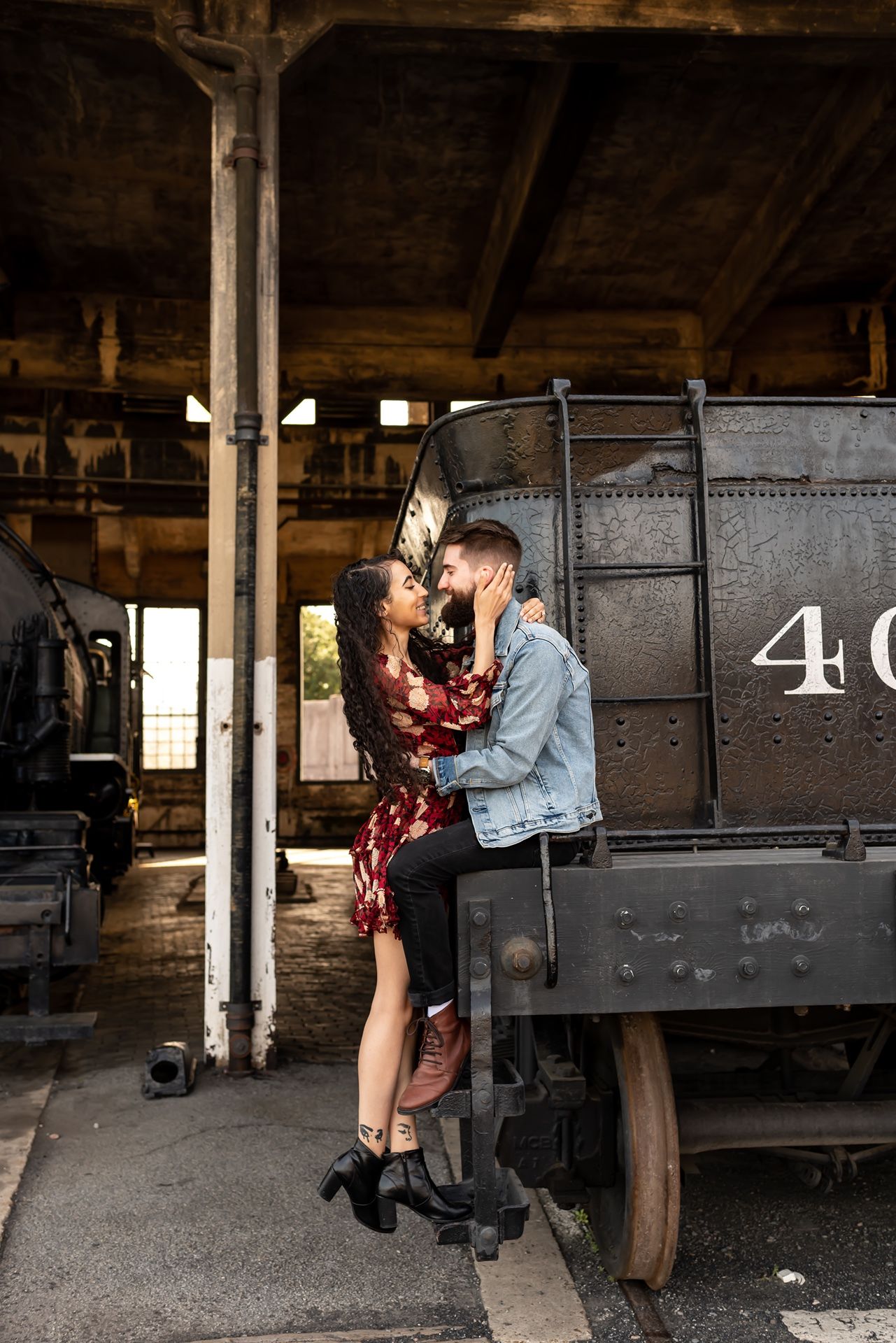 A romantic couple embraces near vintage train cars in a rustic industrial setting, creating a moody atmosphere.