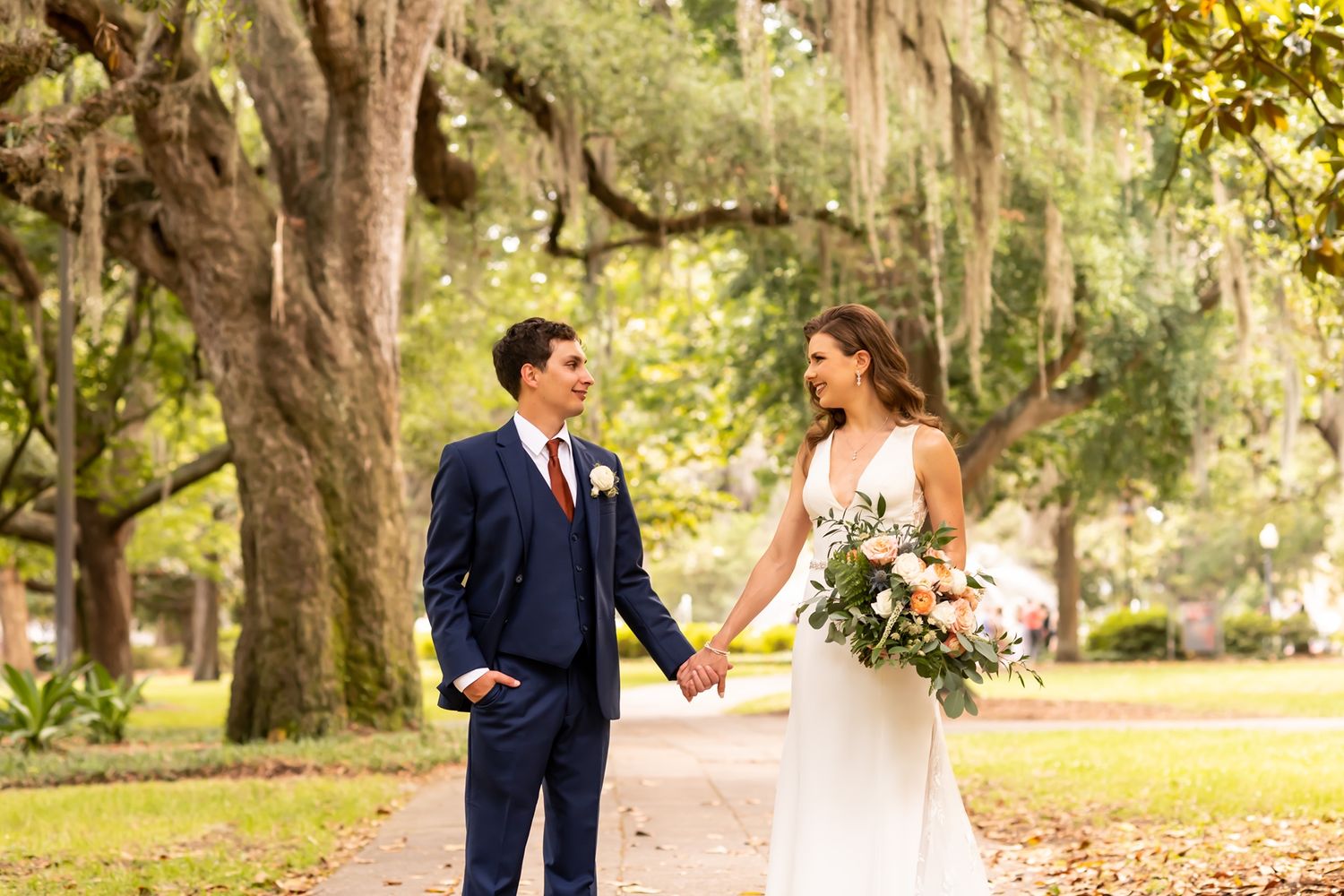 Couple stands together on a scenic tree-lined path with Spanish moss hanging from oak branches in a sunlit garden setting.