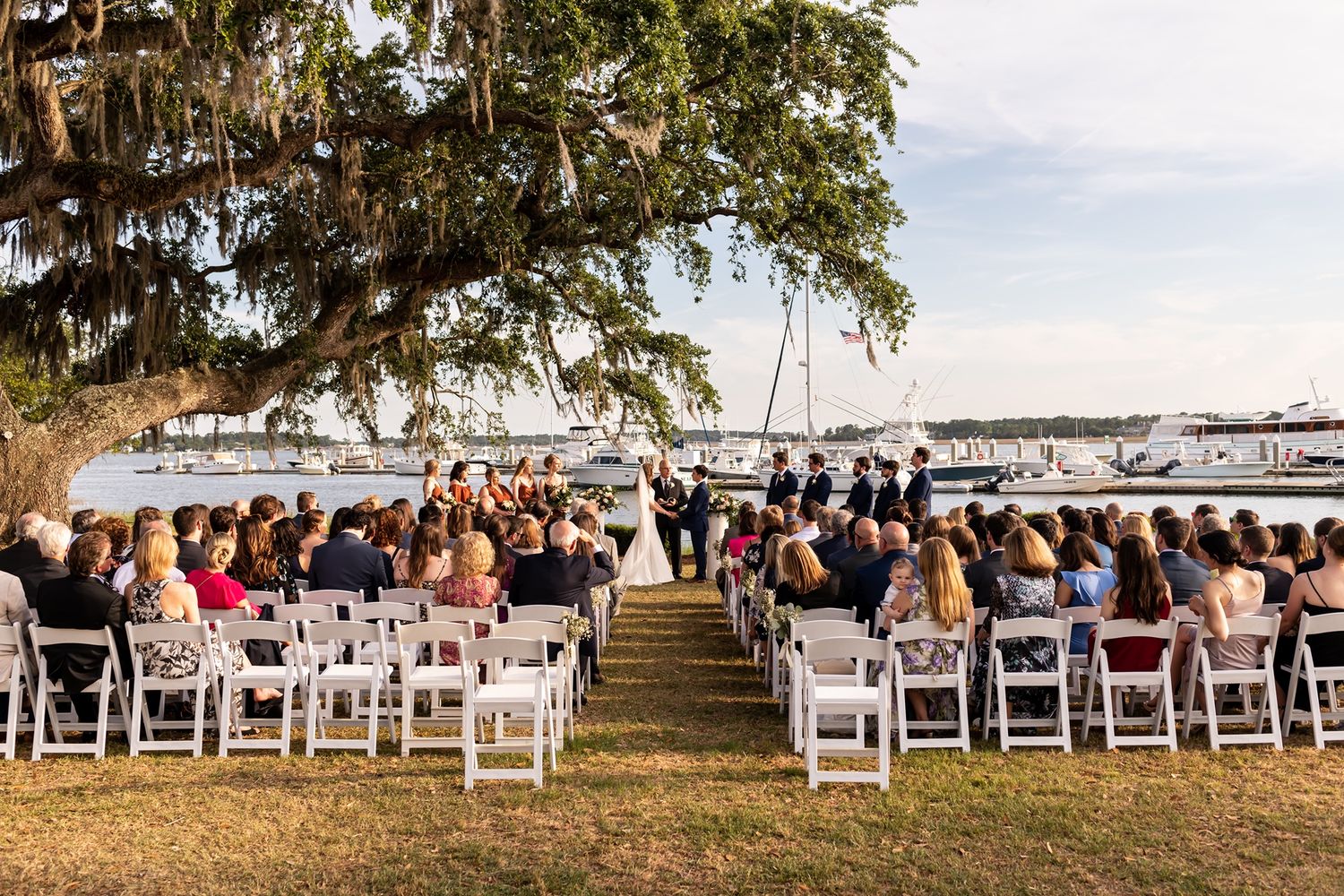 Outdoor wedding ceremony setup with white chairs on grass under Spanish moss tree overlooking waterfront marina.