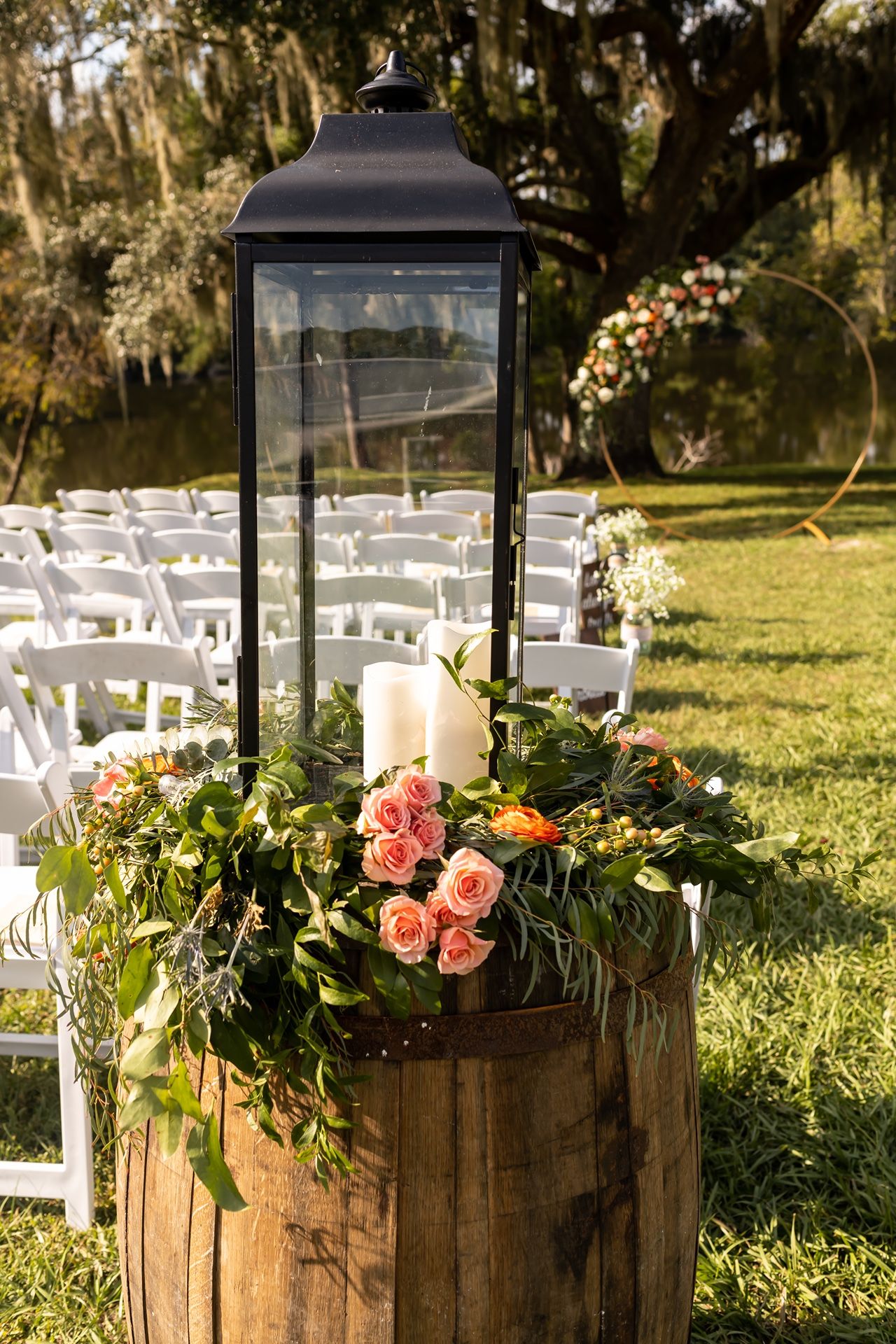 Rustic wooden barrels decorated with pink roses and lanterns line a garden wedding aisle with white chairs.