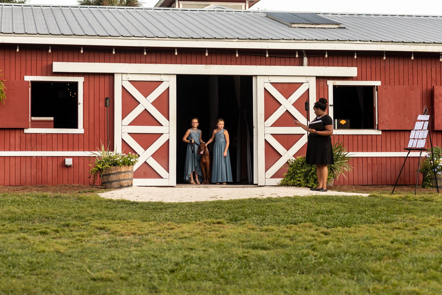 People stand in the doorway of a classic red barn with white X-trimmed doors on a grassy lawn.