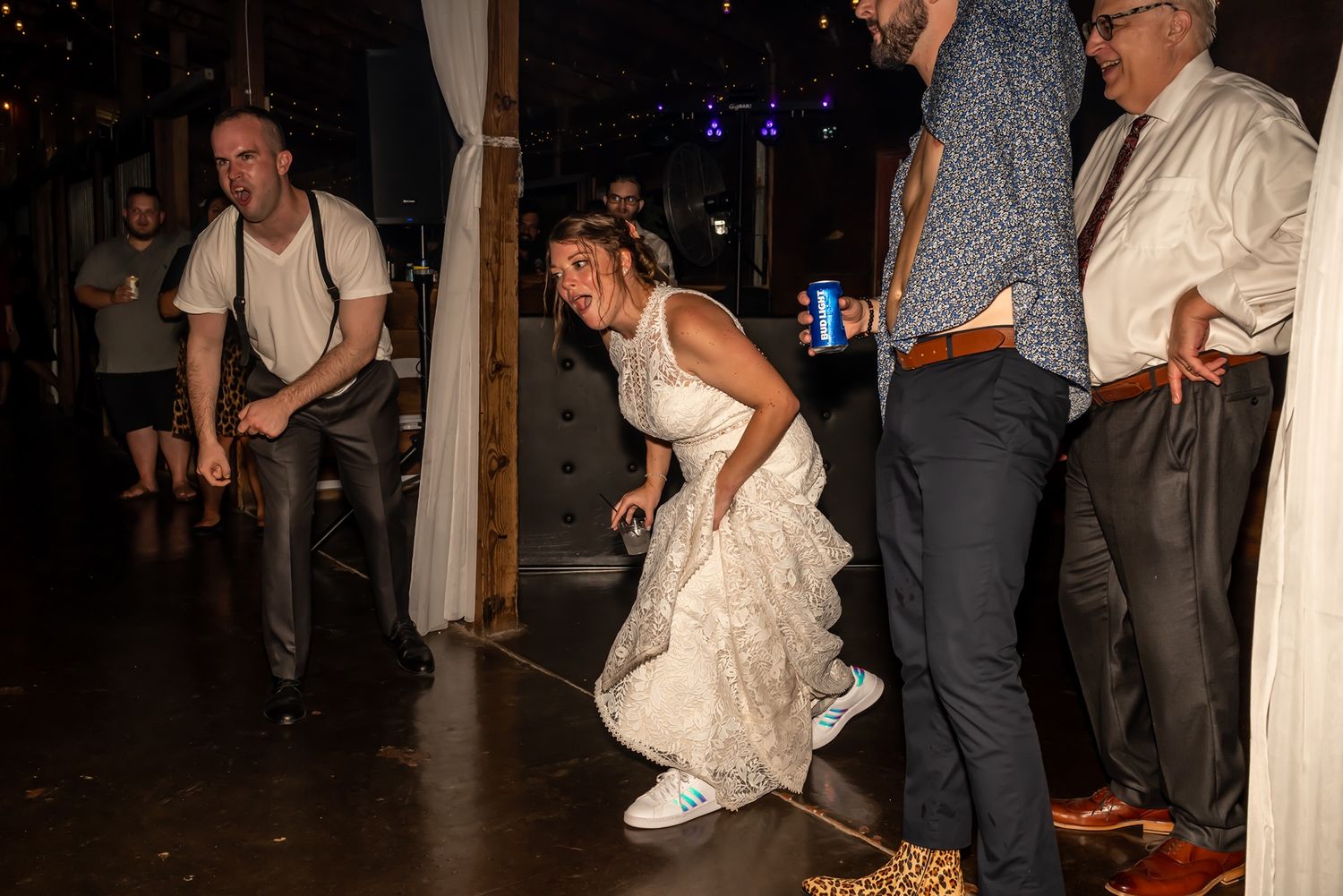 Wedding guests dance and celebrate at a dimly lit reception venue with uplighting and decorative elements.
