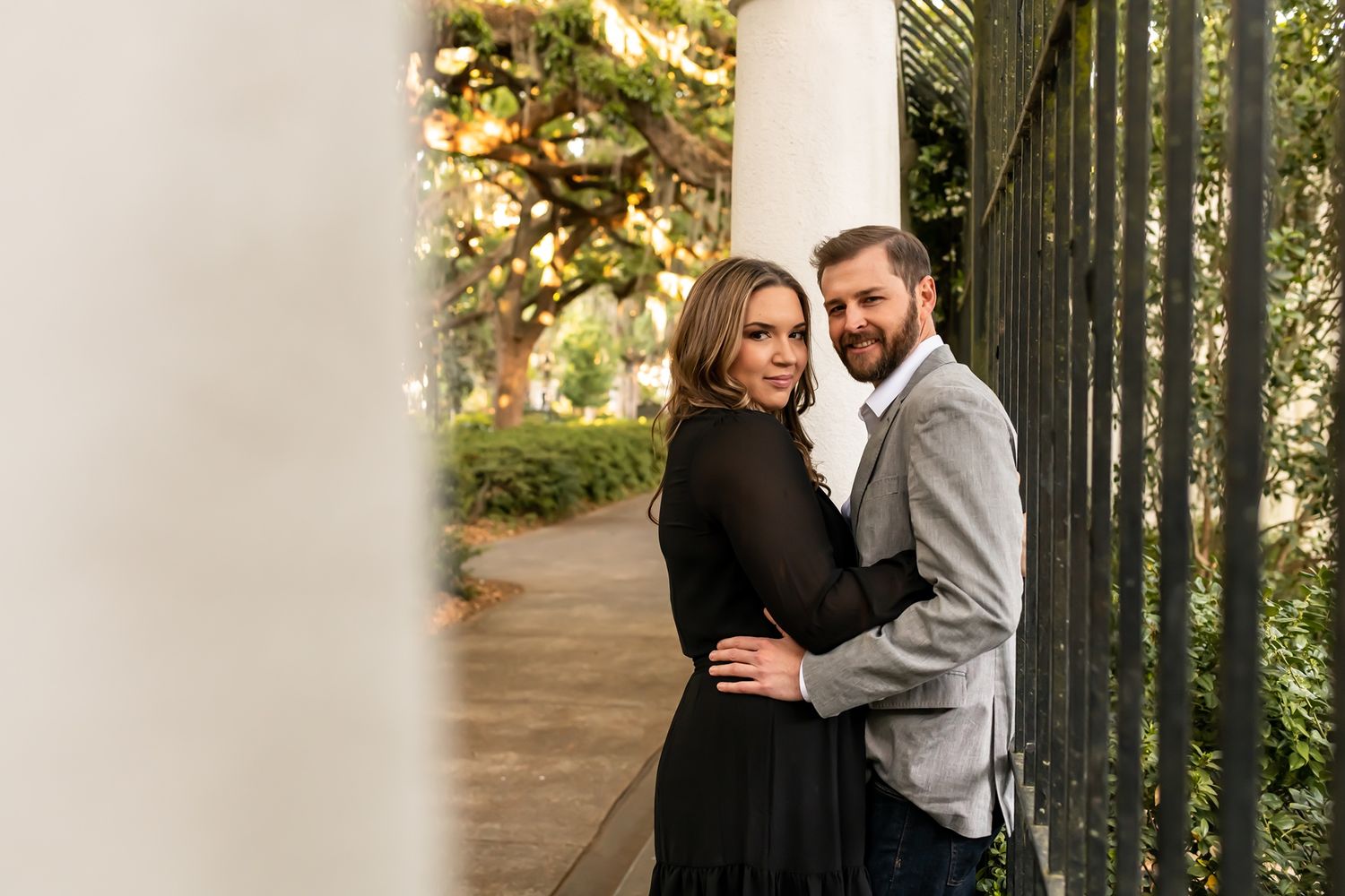 A romantic couple embraces near a white column and iron railing in an outdoor garden setting at sunset.