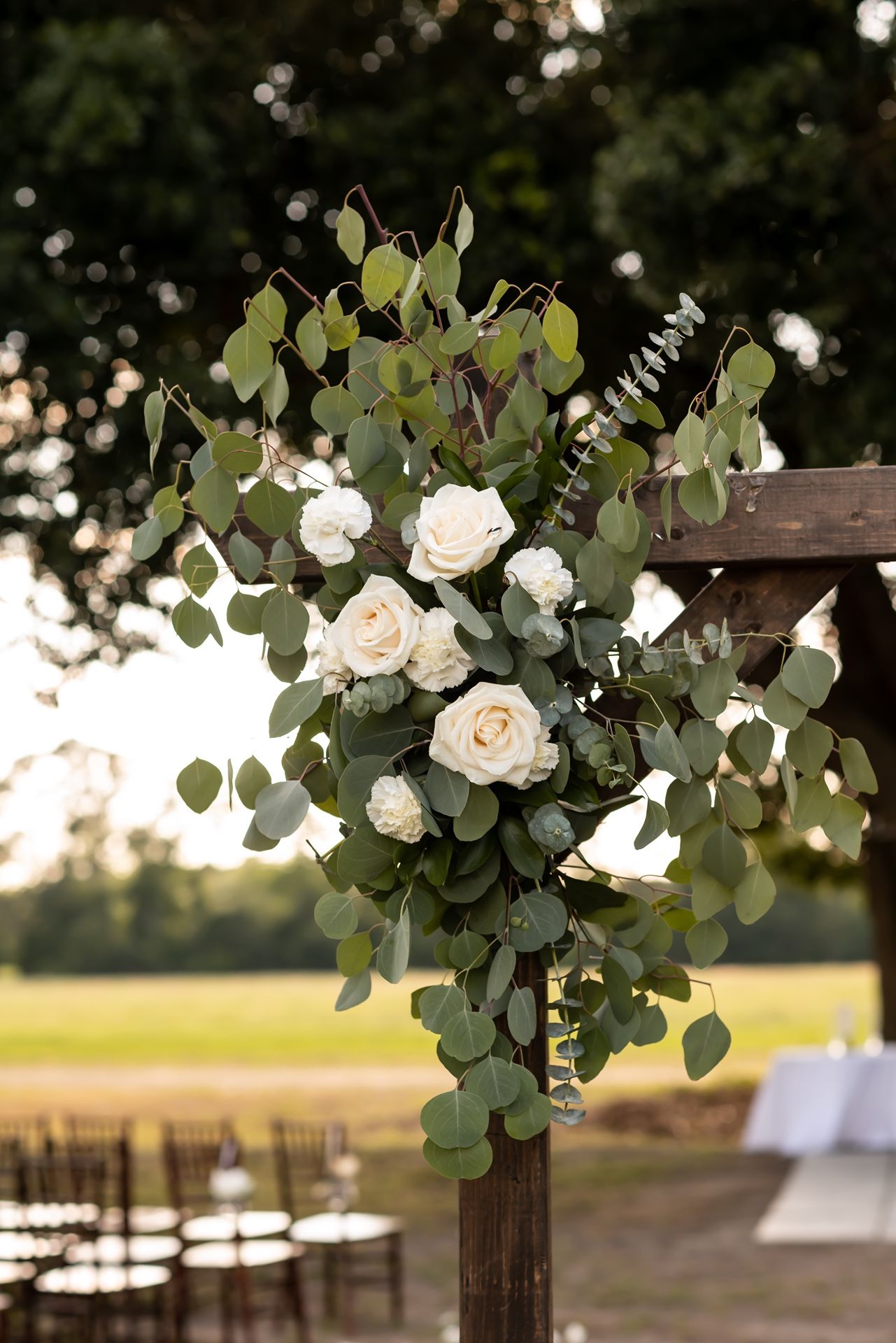 An elegant floral arrangement of white roses and eucalyptus leaves adorns a wooden post in an outdoor wedding setting at sunset.