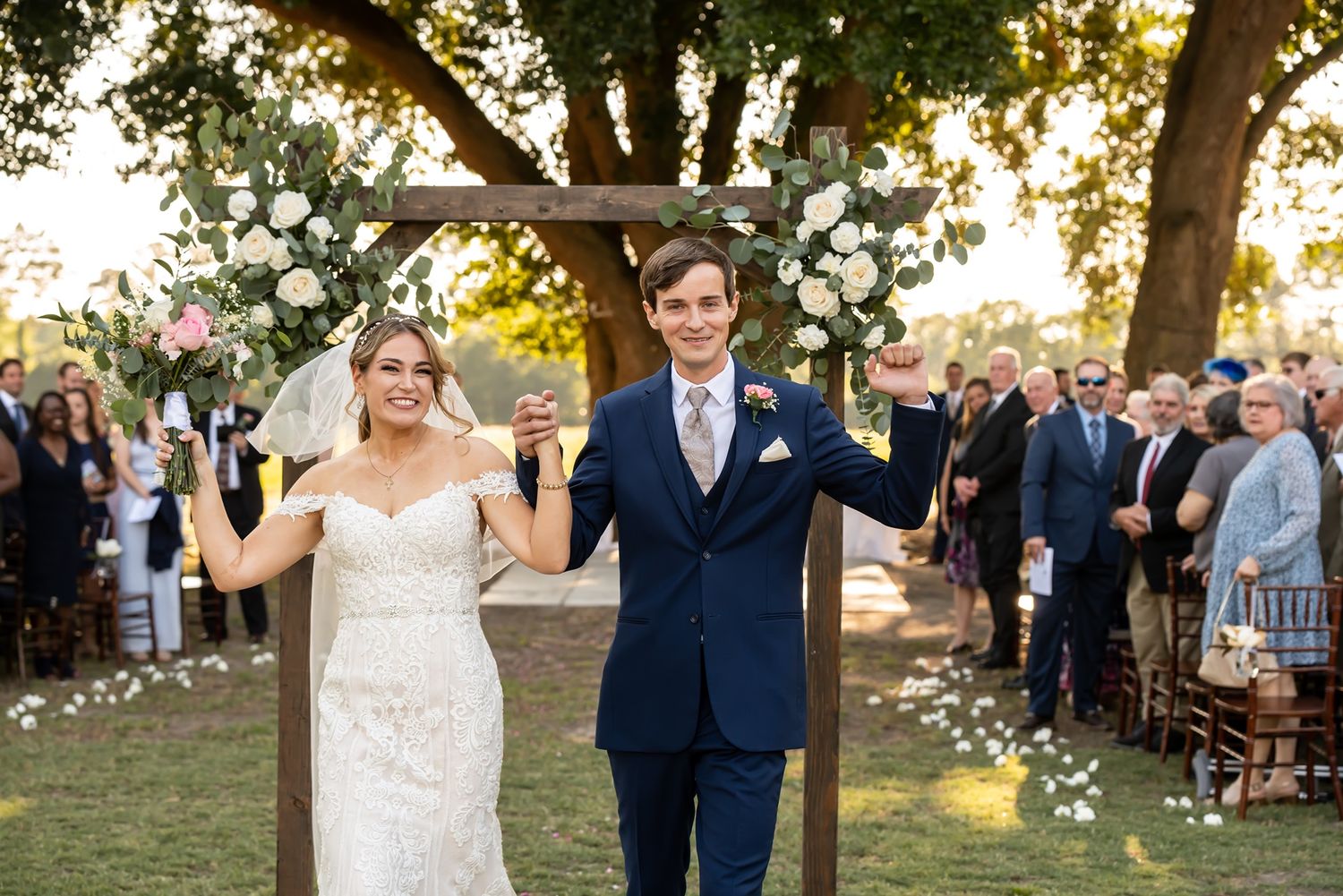A couple raises their hands in celebration under a floral wedding arch during an outdoor garden ceremony with guests watching.