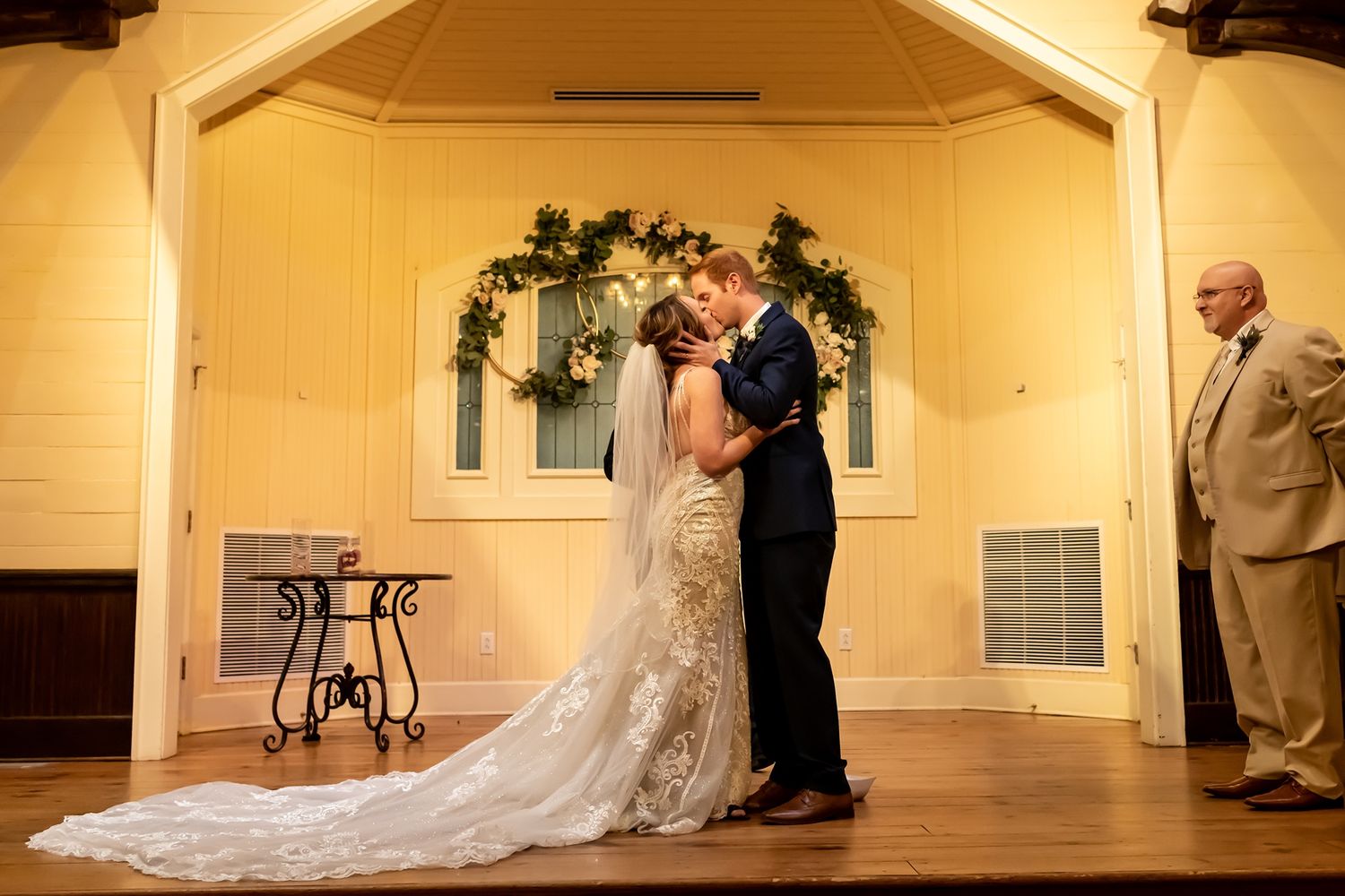 A couple shares their first dance in a warm-lit chapel with yellow walls and a decorative wreath.