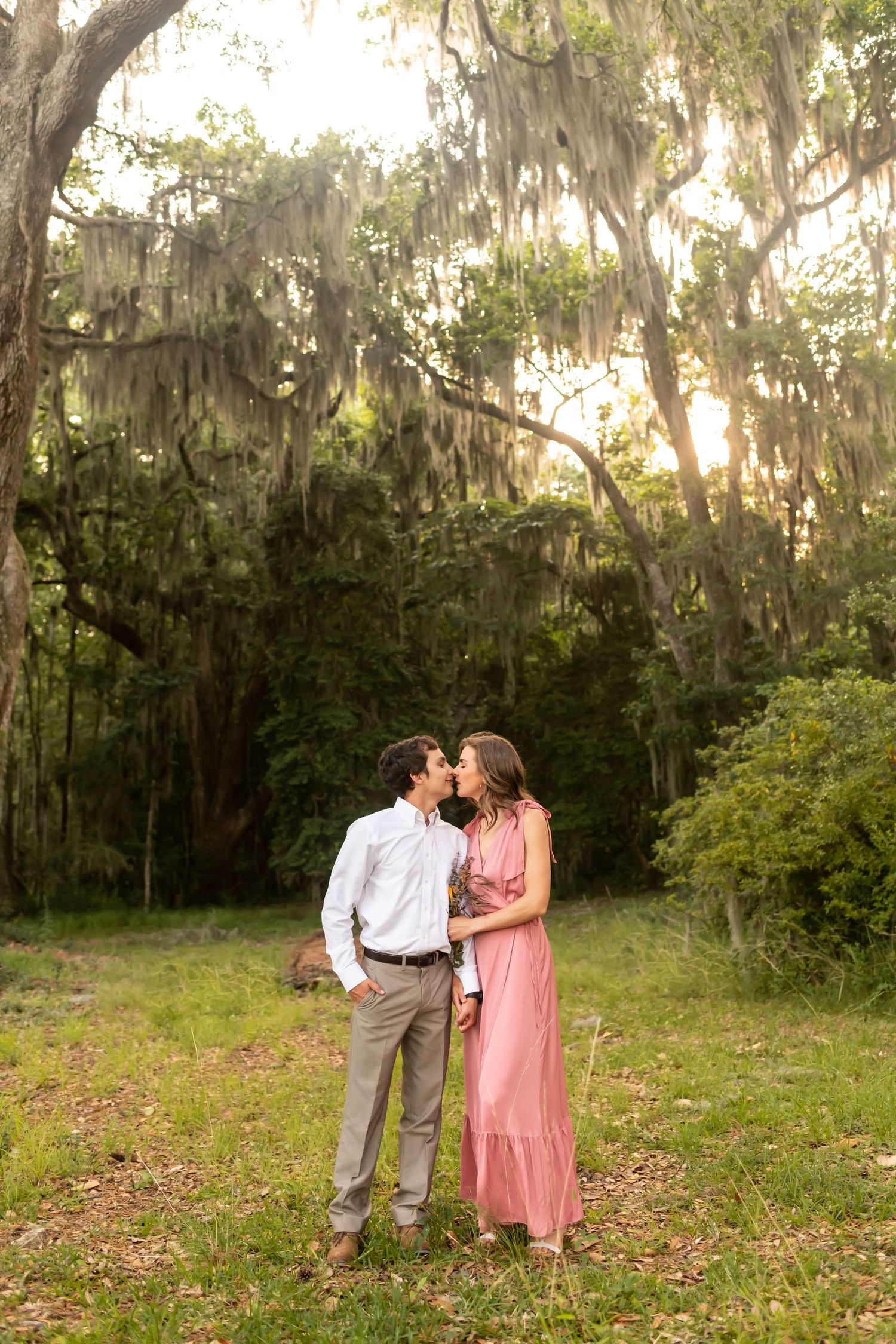 A couple embraces in a dreamy garden setting with Spanish moss draped oak trees in the background at sunset.