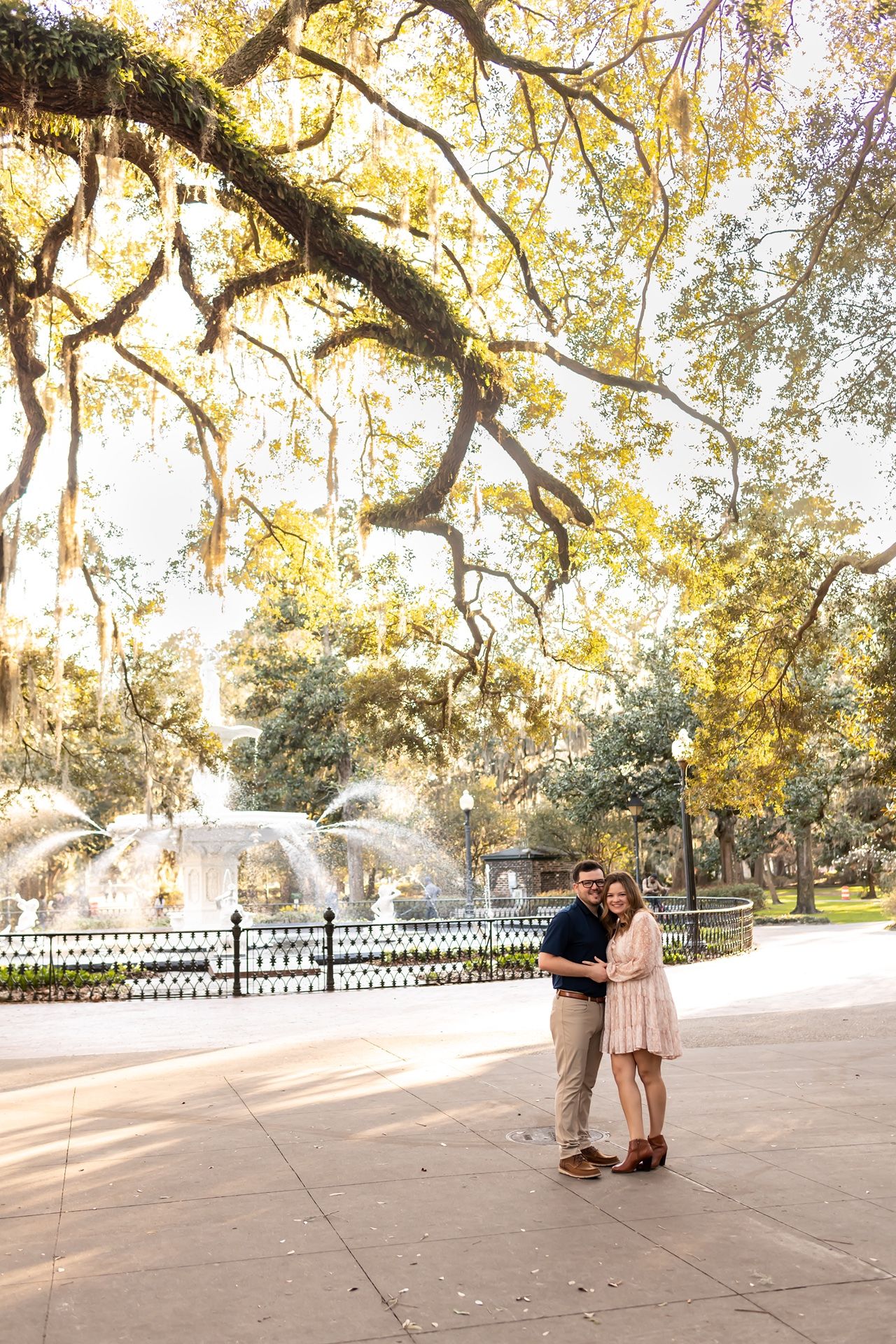 A couple embraces in front of a fountain under Spanish moss-draped trees in a sun-lit park setting.