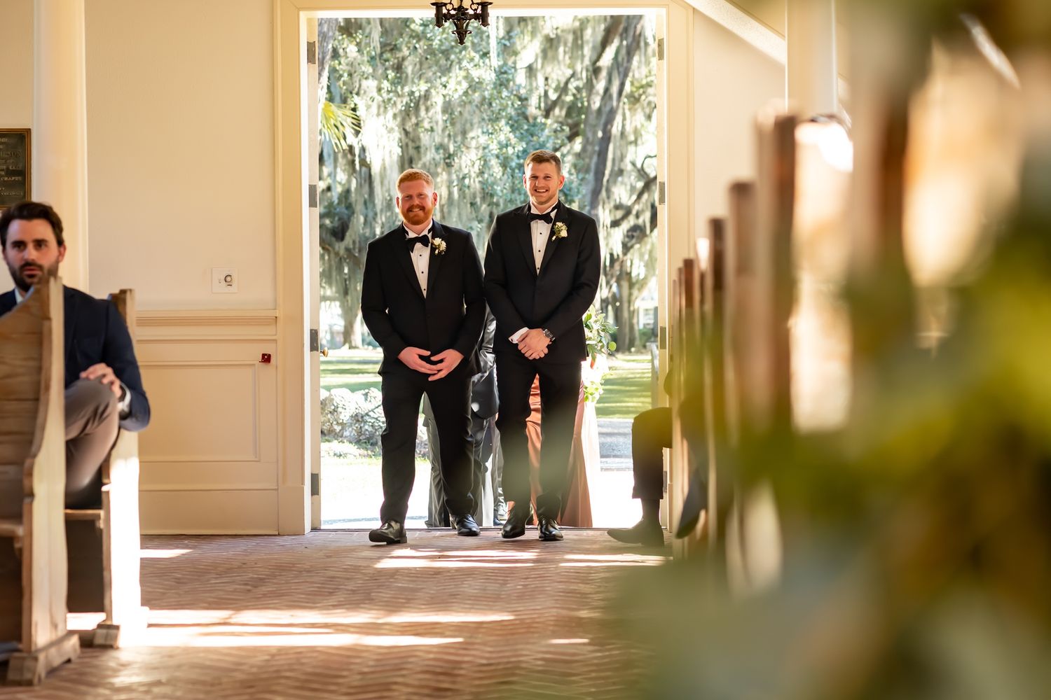 Two people in black tuxedos standing in a bright doorway with hanging Spanish moss visible outside.