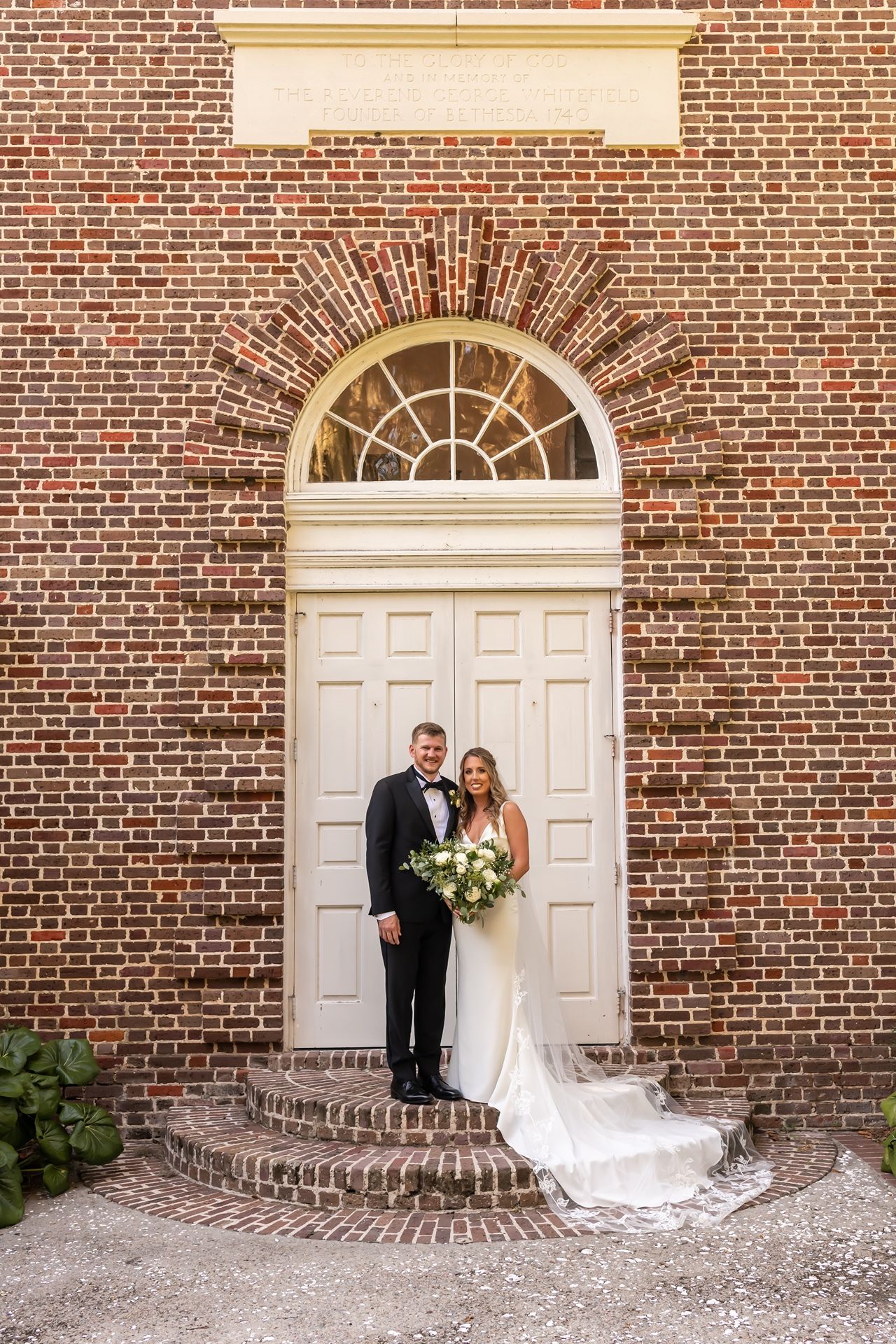 Elegant couple poses in front of historic brick church entrance with white door and arched window, surrounded by traditional architecture.