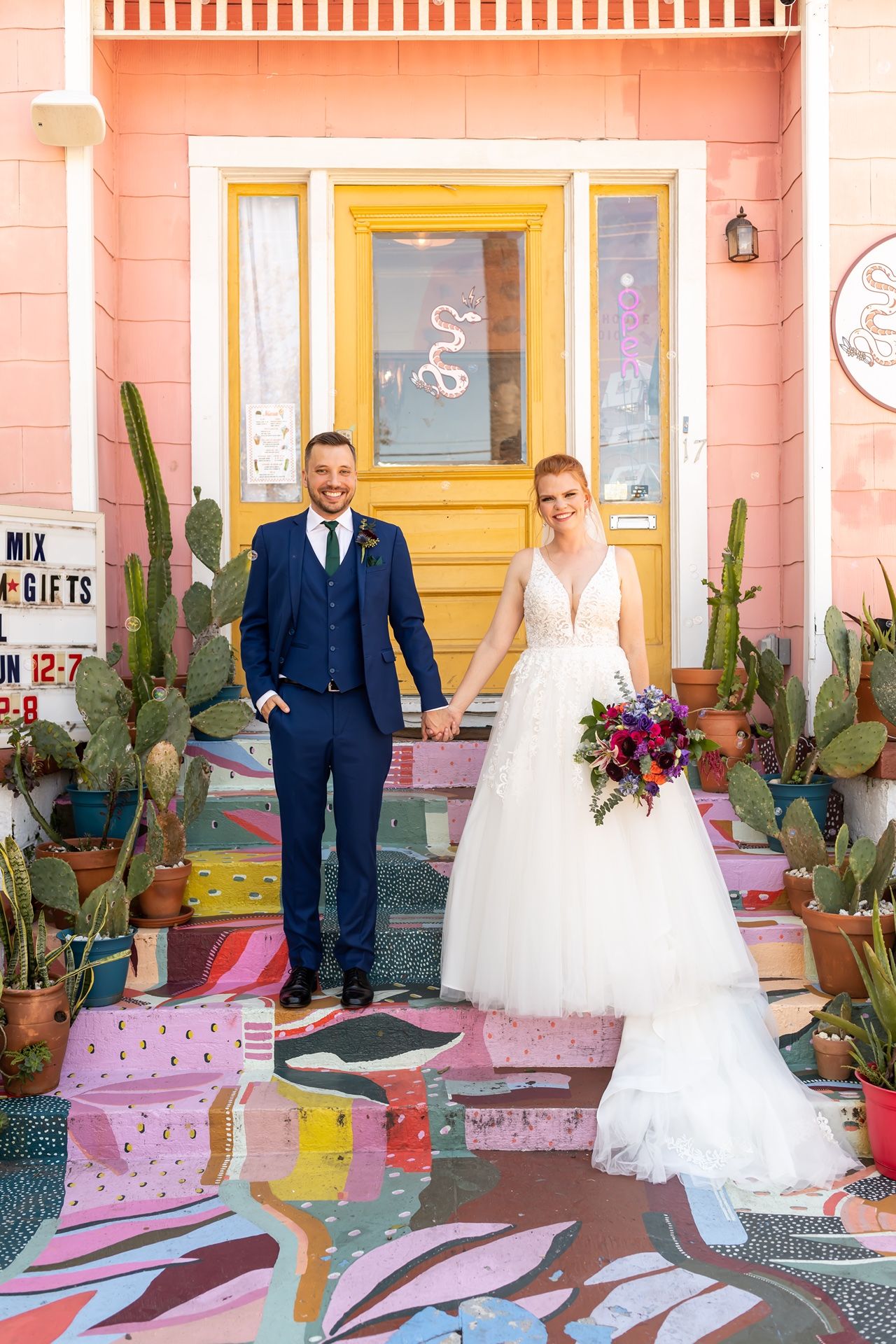 A pink cactus-lined facade with white railing, yellow door and colorful wedding decor creates a vibrant photo backdrop.