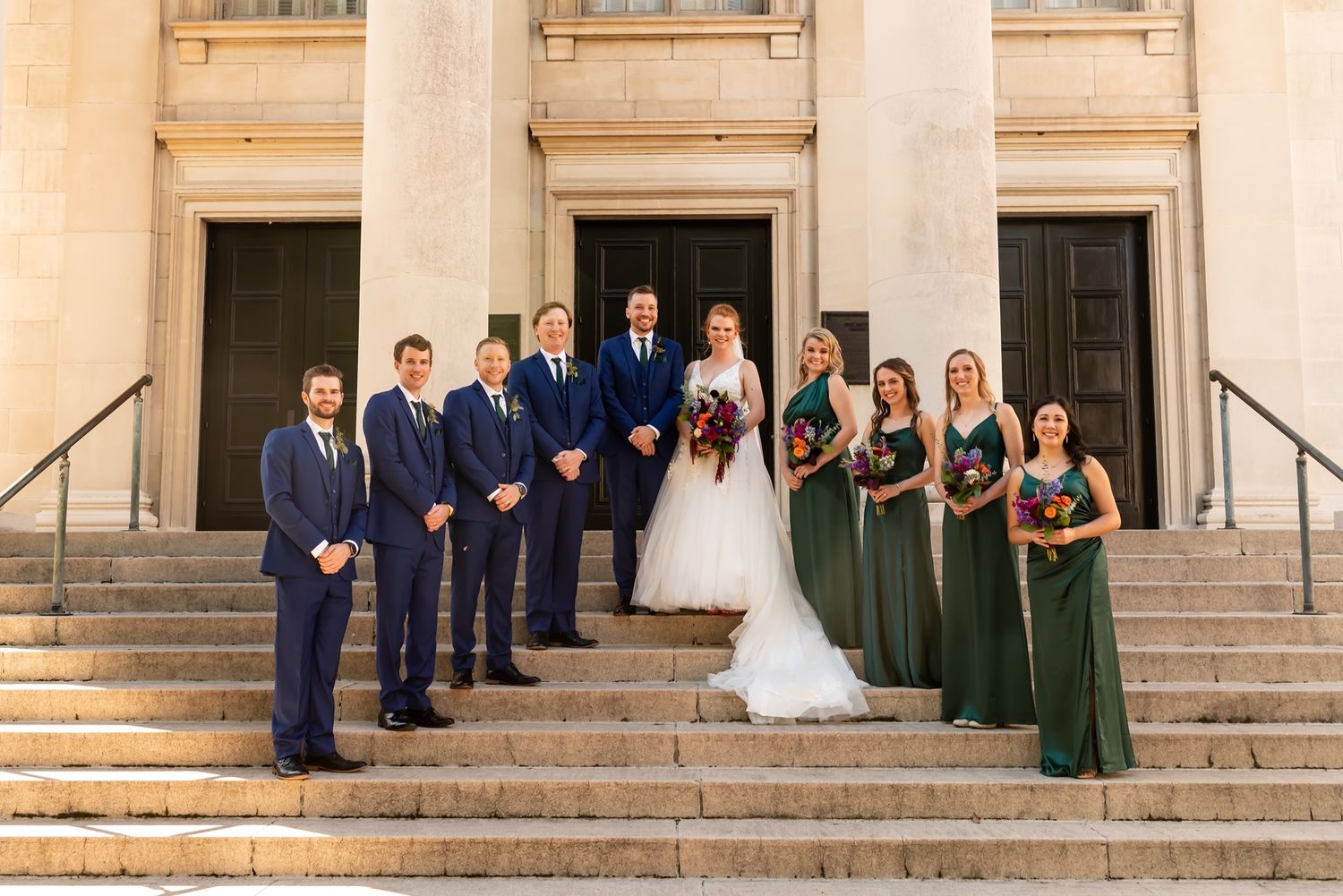 Wedding party in navy suits and green dresses poses on grand stone steps of historic building entrance.