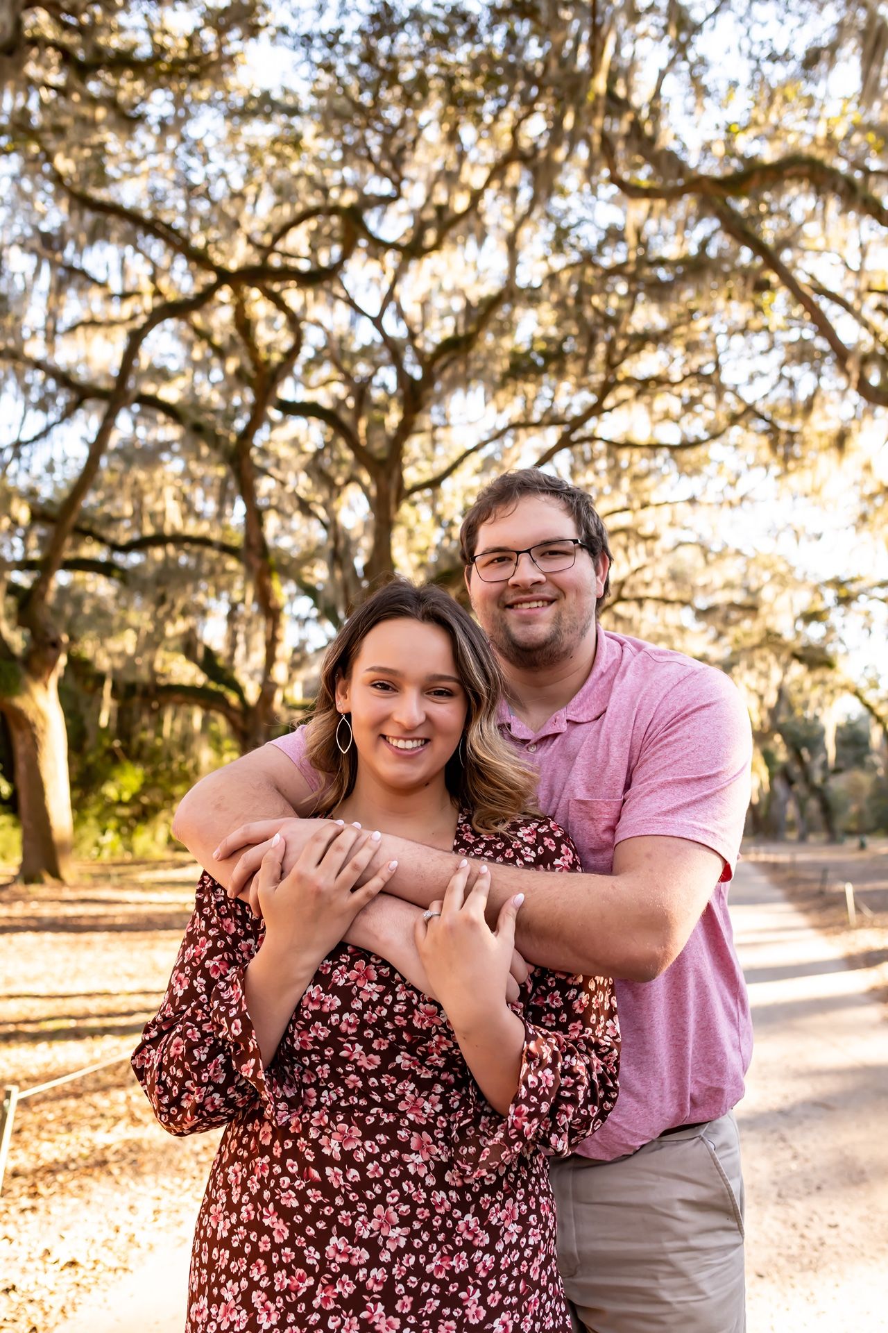 A couple shares a warm embrace under moss-draped oak trees in a sunlit park during golden hour.