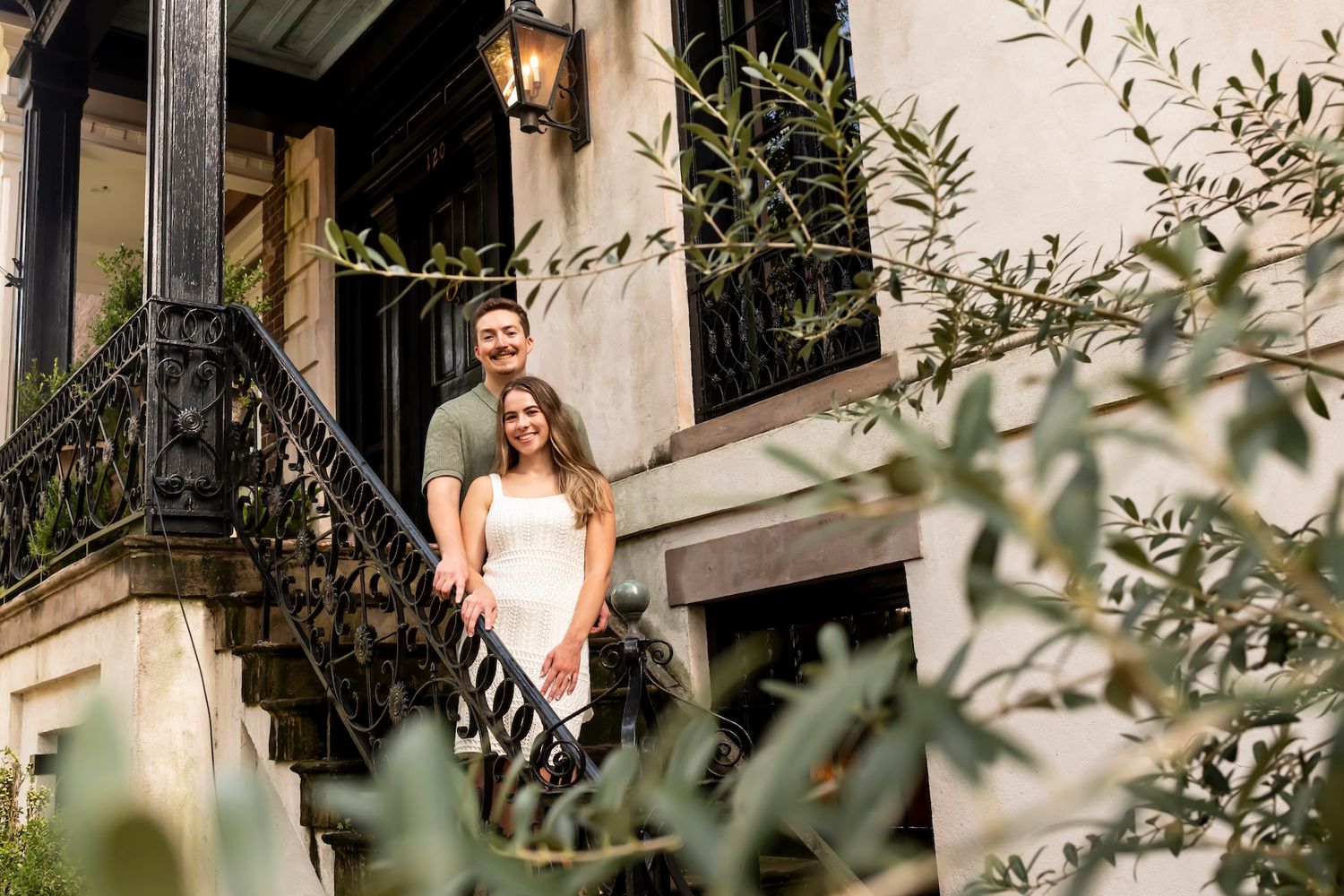 A couple stands on an elegant stone balcony with ornate wrought iron railings surrounded by greenery and olive branches.
