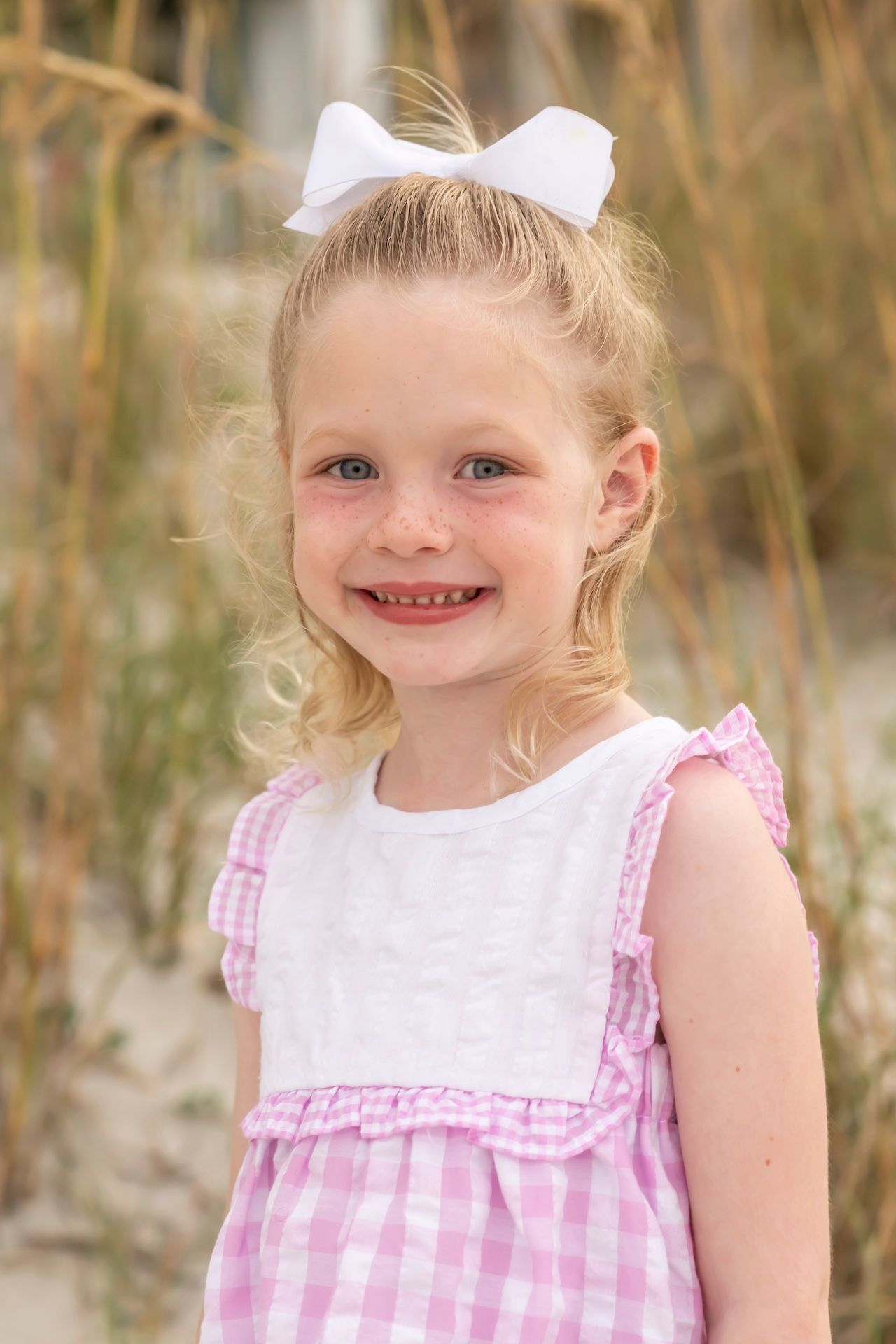 Child in pink and white gingham dress with white bow smiles at camera against natural outdoor setting.