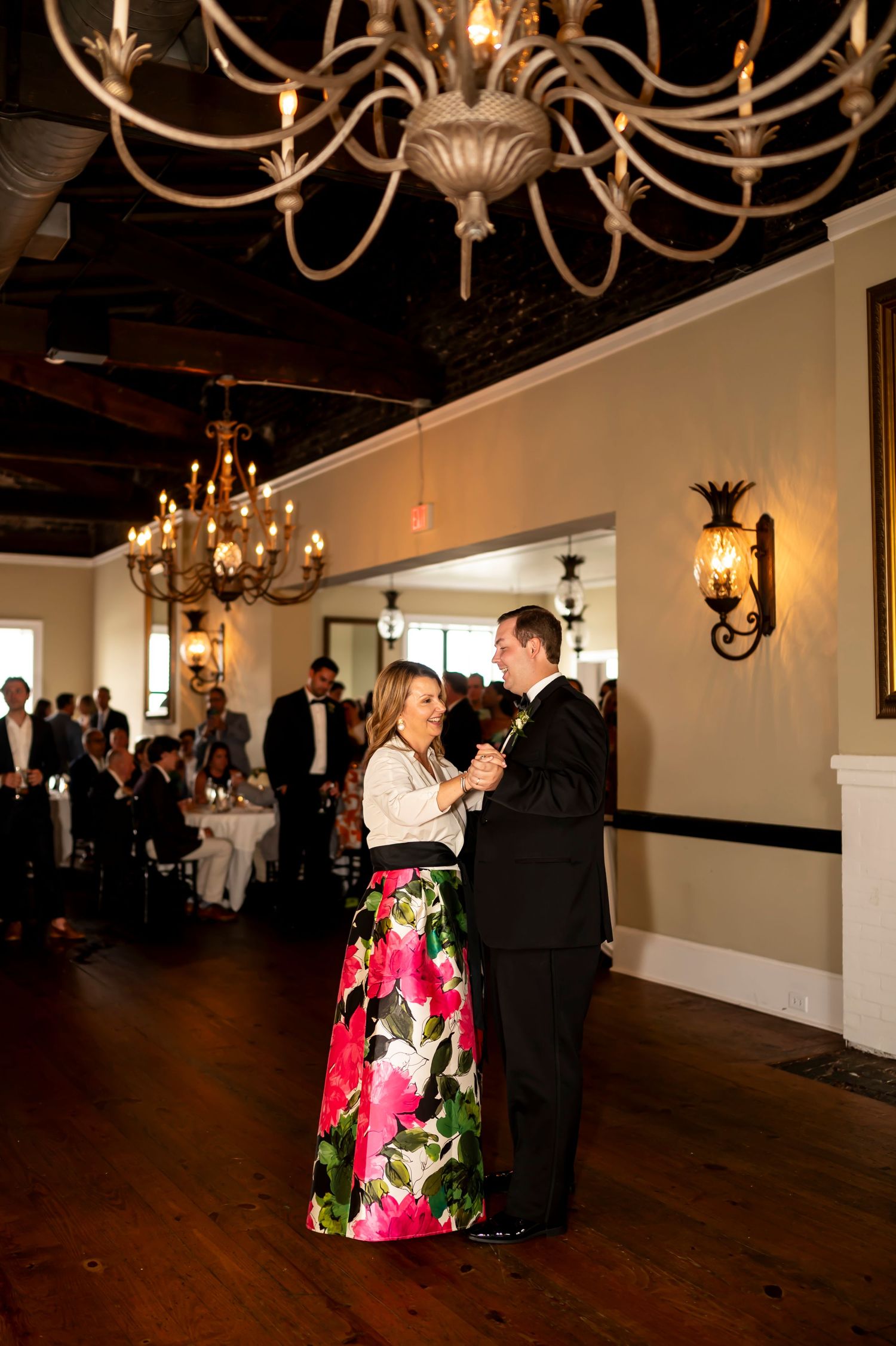 A couple dances together in an elegant reception hall with ornate chandeliers and wall sconces under warm lighting.