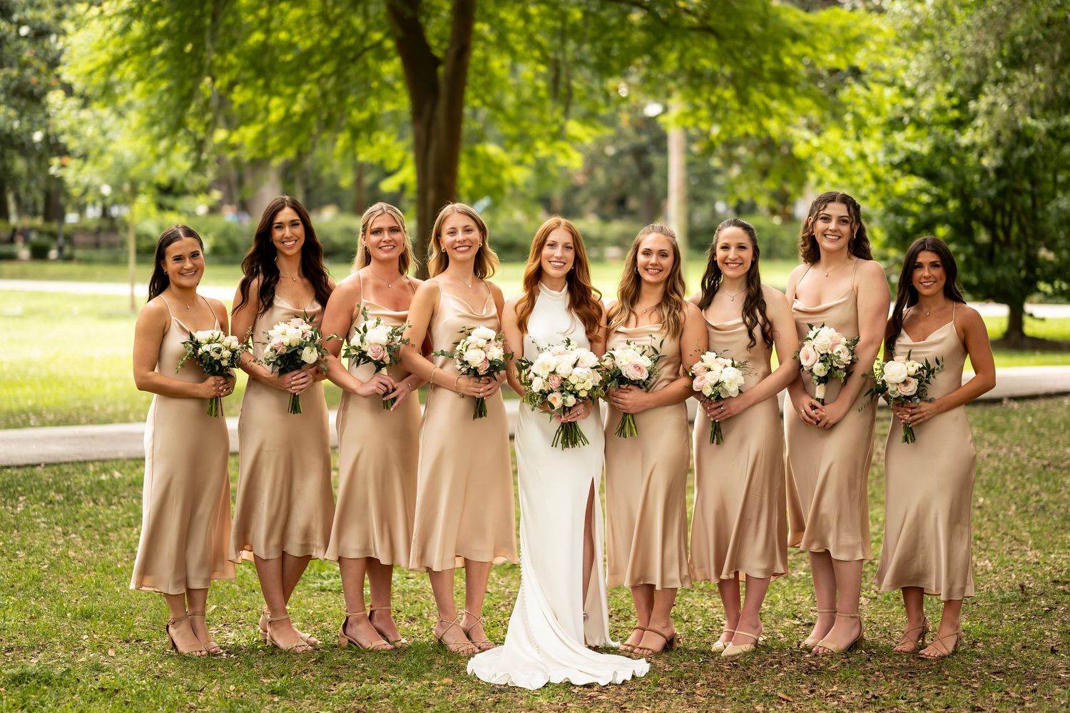 A bridal party stands together in champagne colored dresses holding bouquets in a lush garden setting.