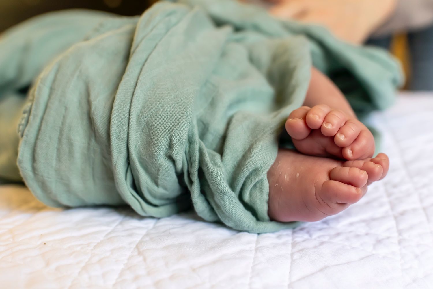 Small baby feet peeking out from under a soft mint green blanket on white bedding.