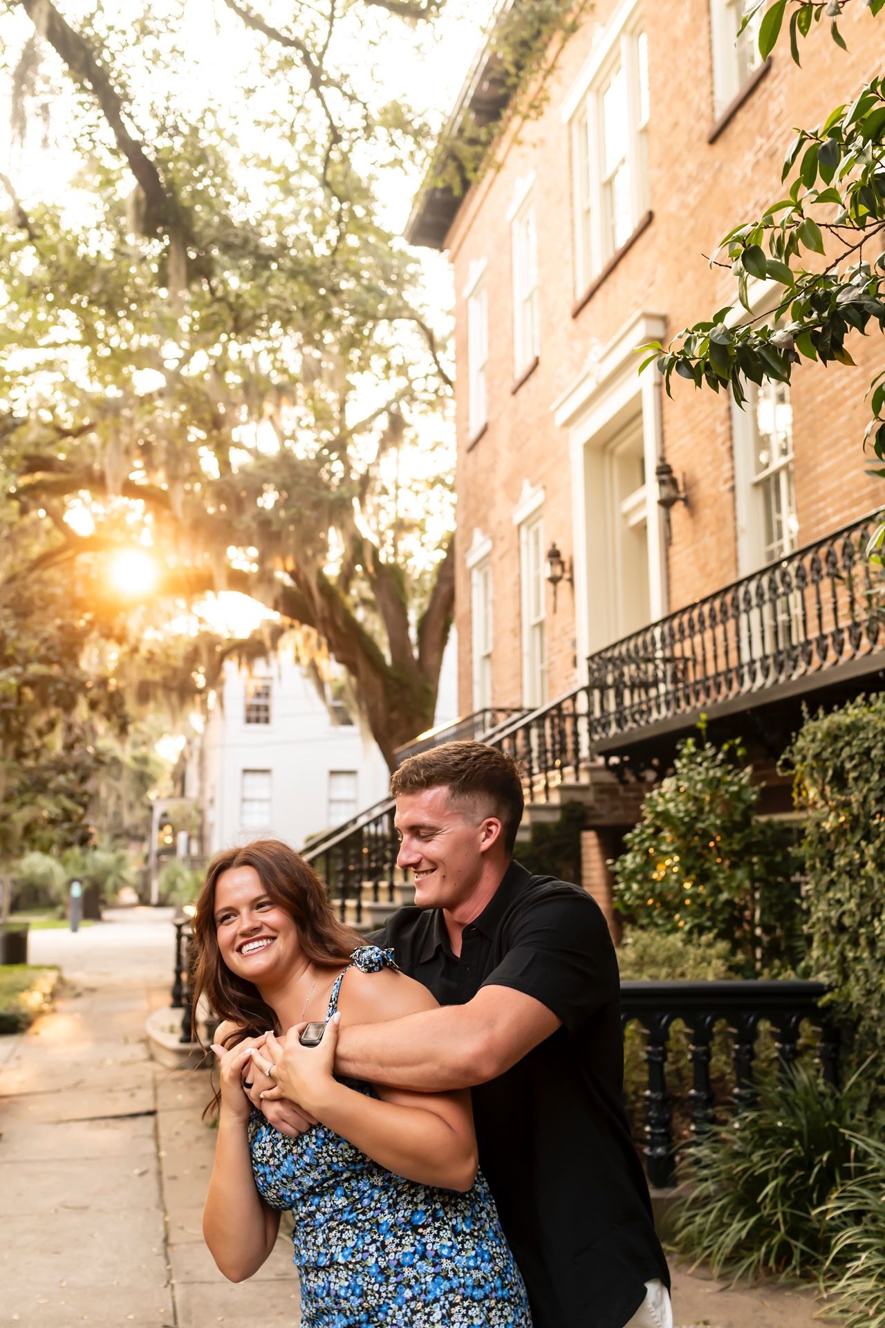 A couple playfully interacts on a tree-lined urban street at sunset in a romantic engagement photo session.