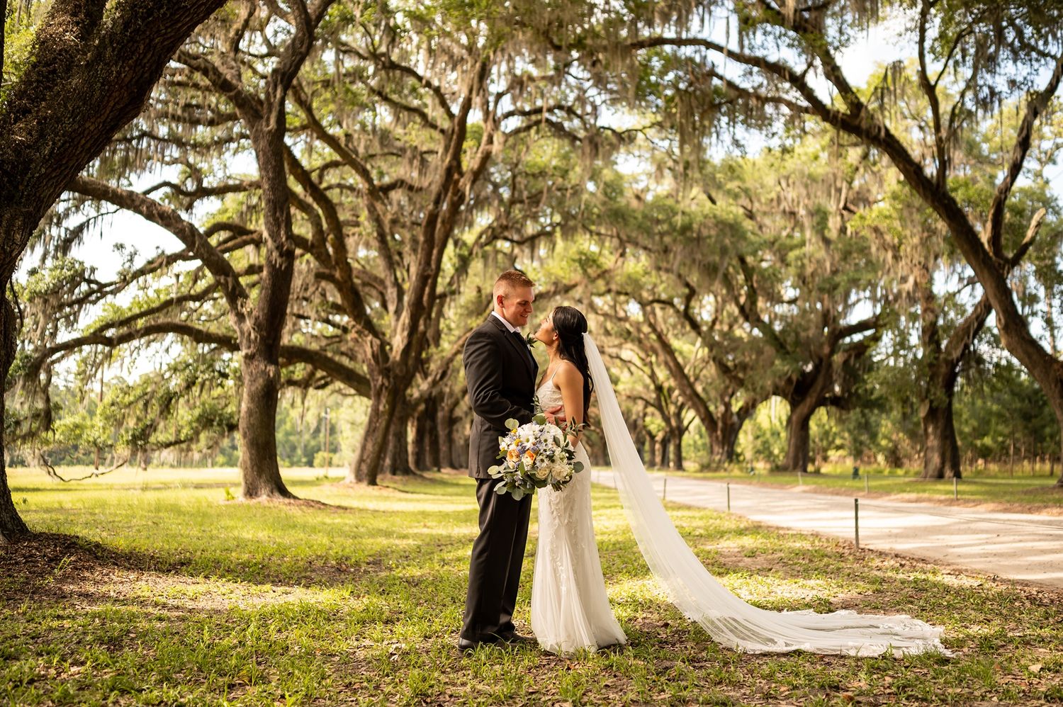A couple stands beneath majestic oak trees draped with Spanish moss during their outdoor wedding ceremony.