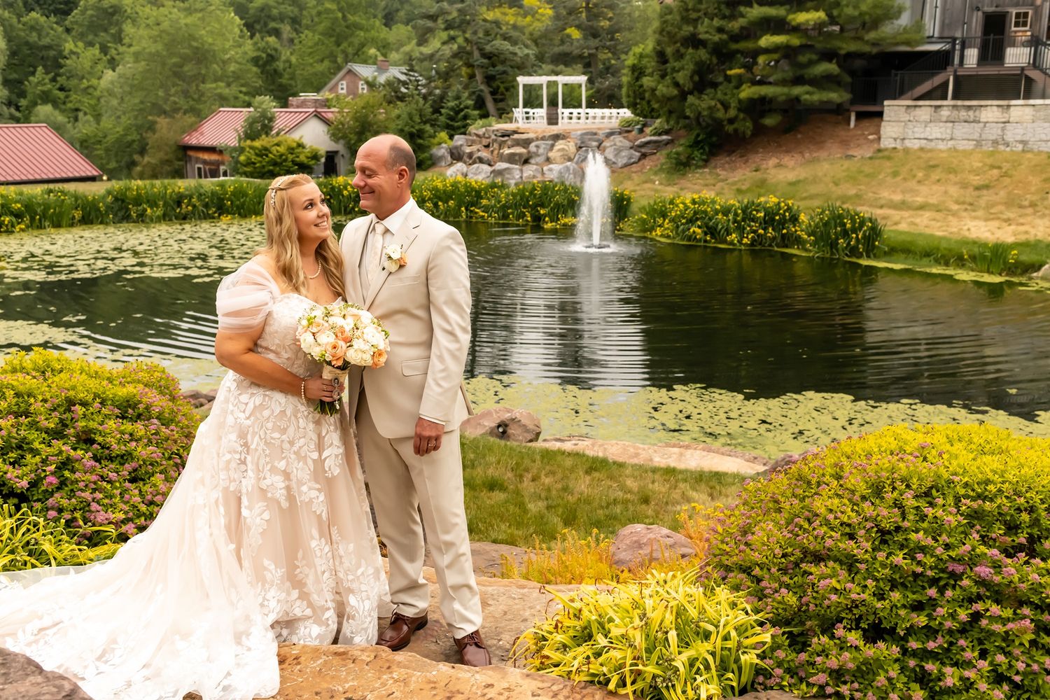 A couple stands together by a pond with a fountain and yellow flowers at an outdoor wedding venue.