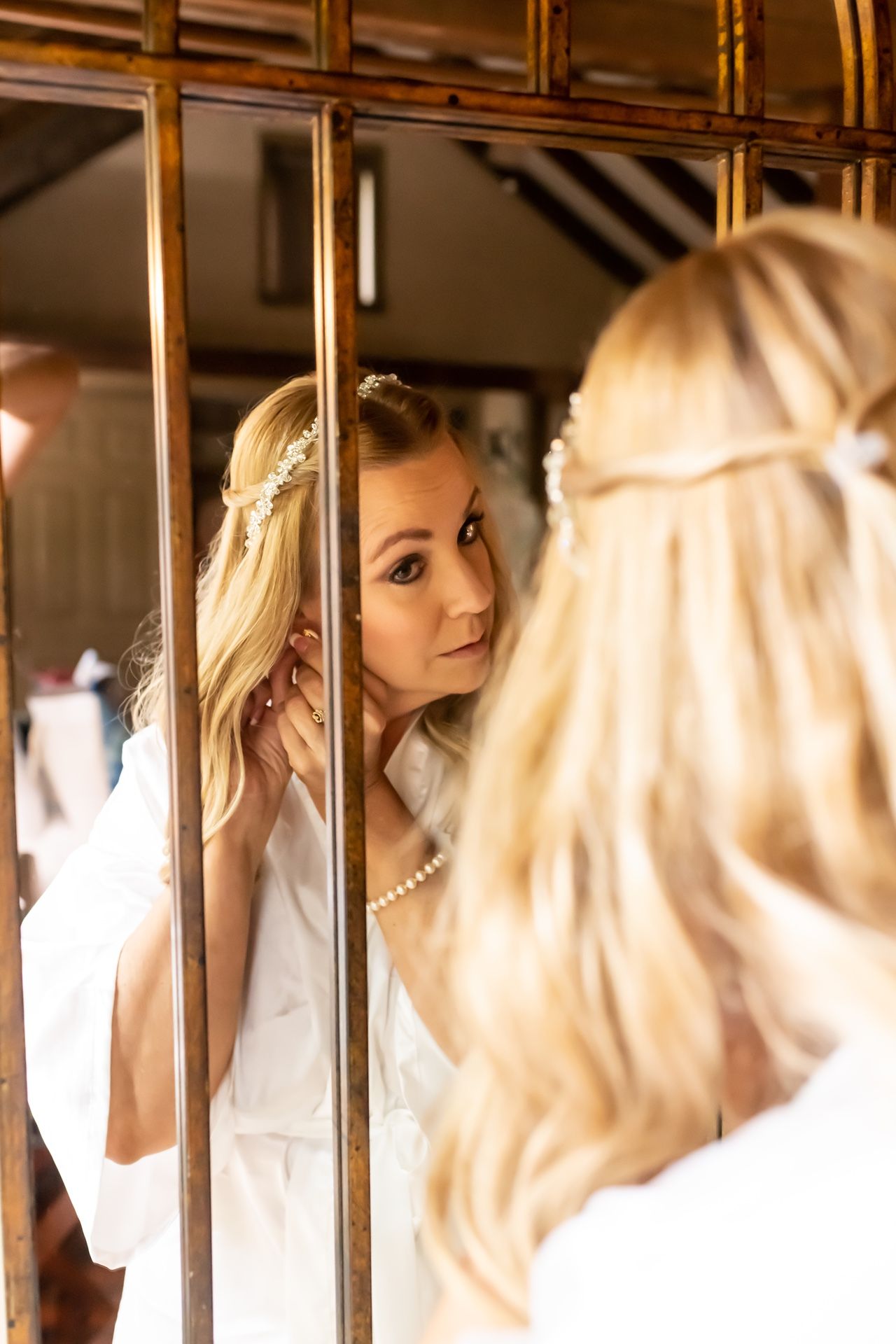 A blonde figure looks into a gold-framed mirror while adjusting jewelry and wearing white clothing.