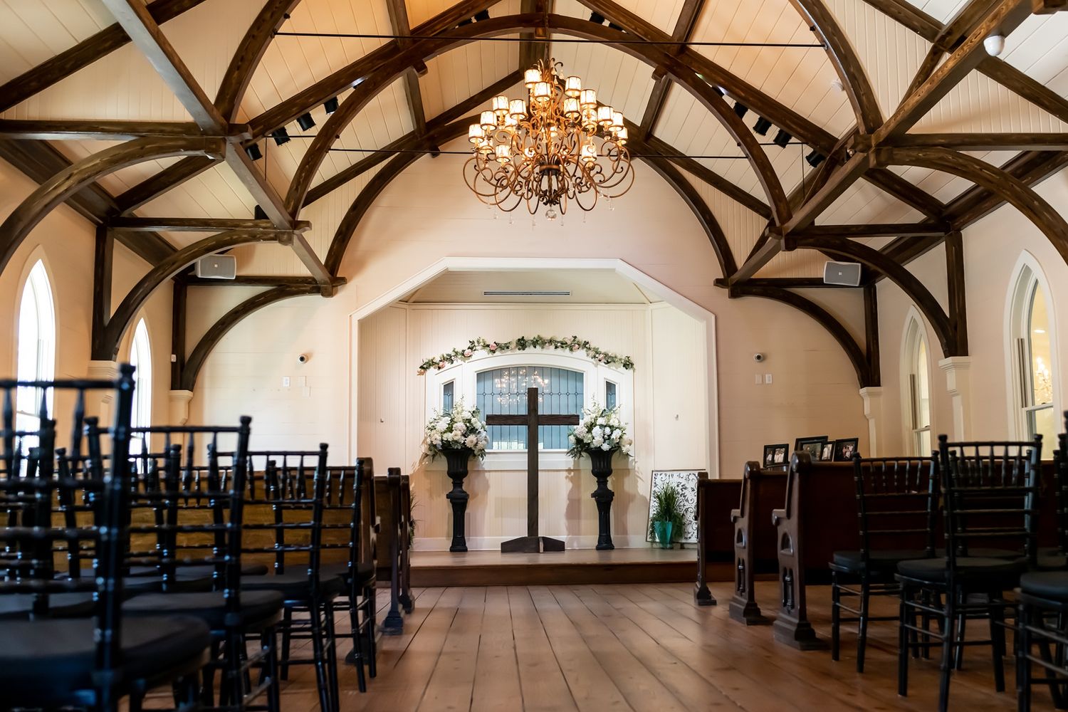 A rustic wedding chapel interior with exposed wooden beams, elegant chandeliers, and rows of dark wooden chairs.