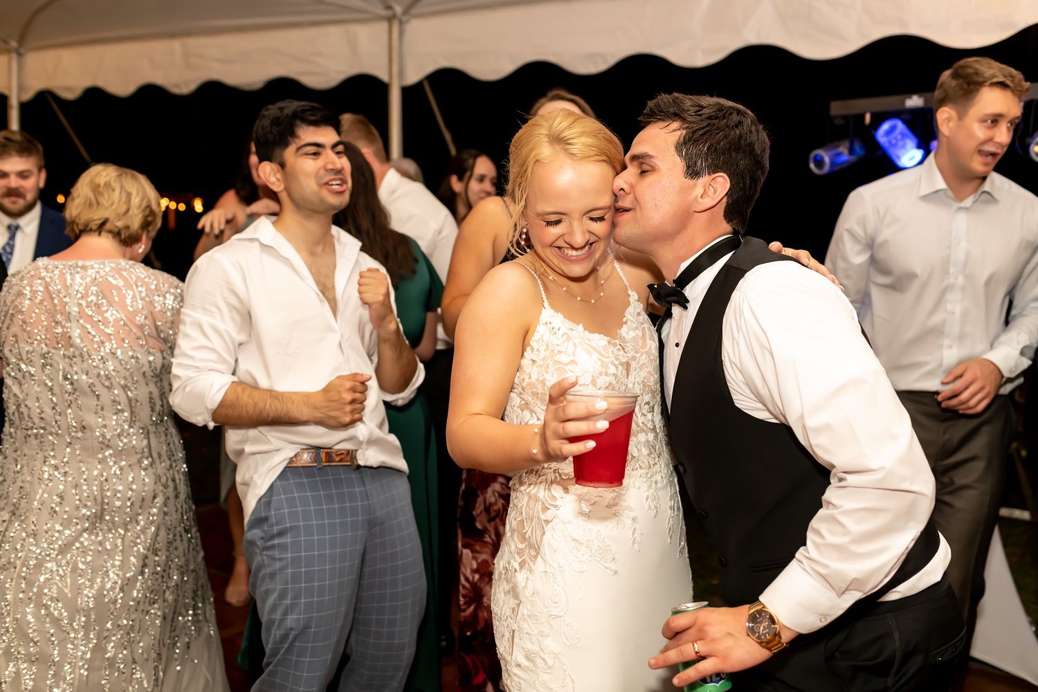 A bride holding a red drink celebrates with wedding guests under a white tent during the reception.