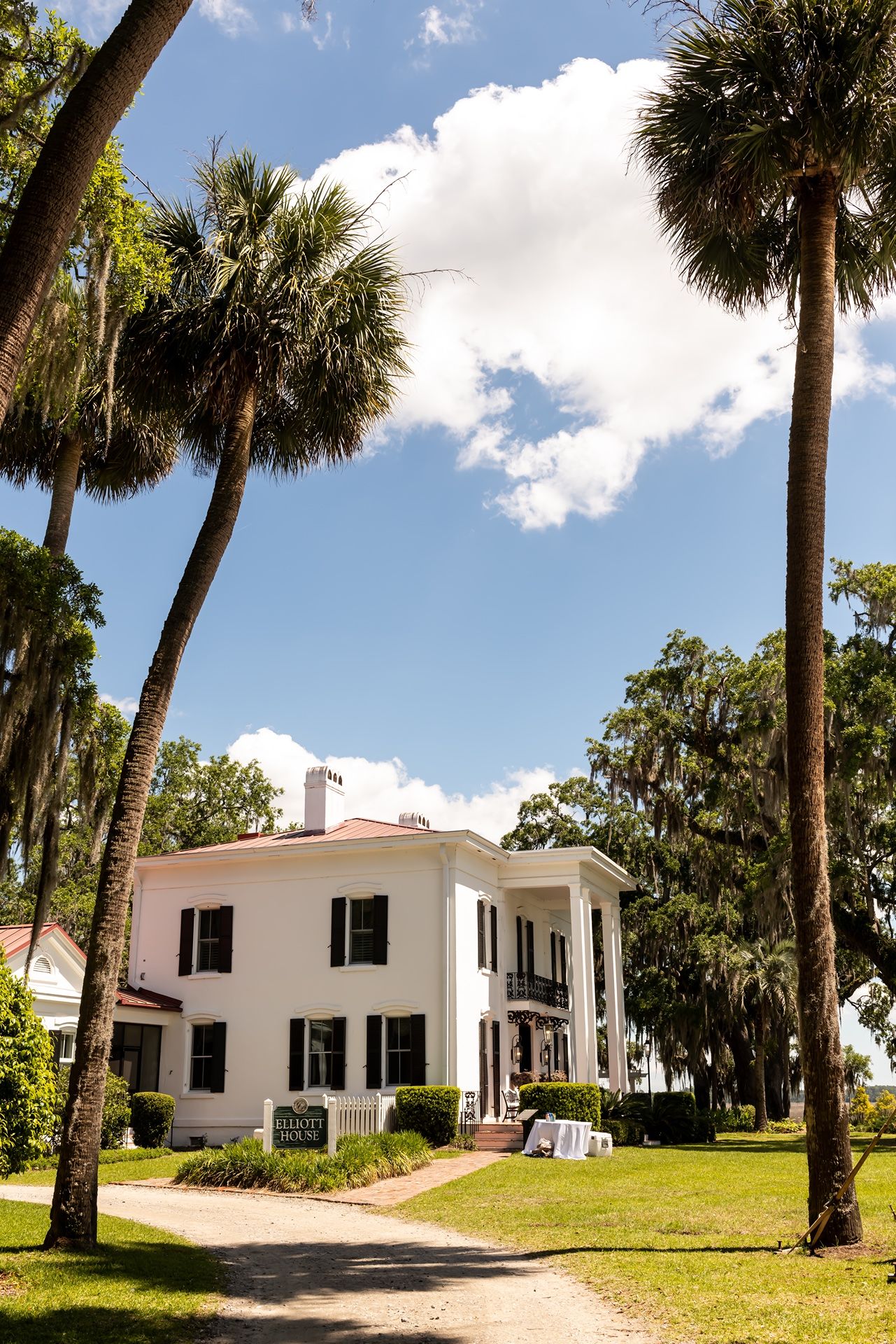 A white two-story Southern plantation-style home with tall palm trees and Spanish moss in the background under blue skies.