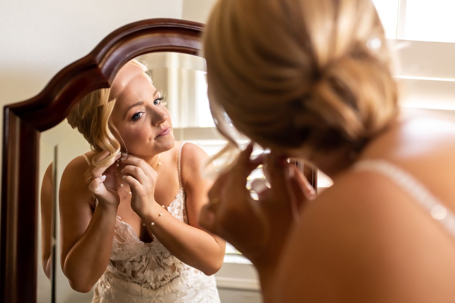 A bride in an elegant white dress puts on earrings while looking in an antique mirror during wedding preparations.