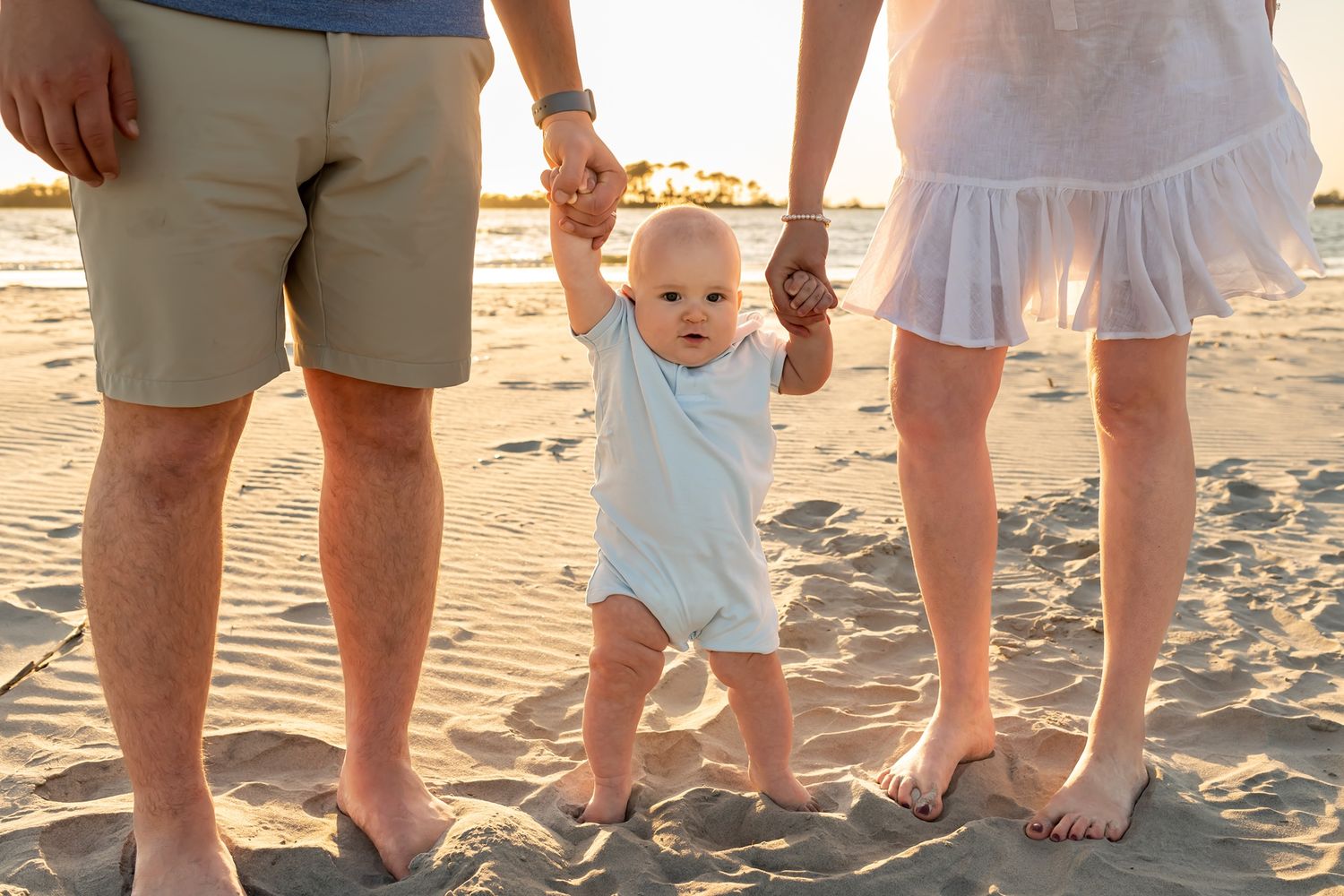 Parents holding hands with toddler taking first steps on sandy beach at sunset.