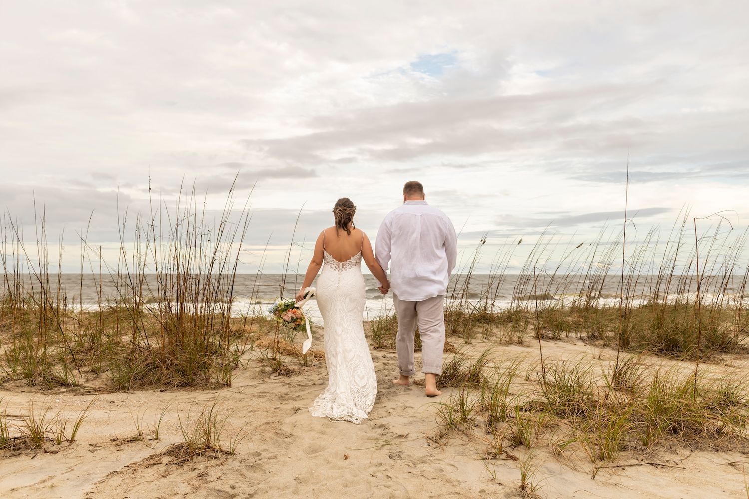 A couple walks hand in hand along a sandy beach path through sea oats at sunset.