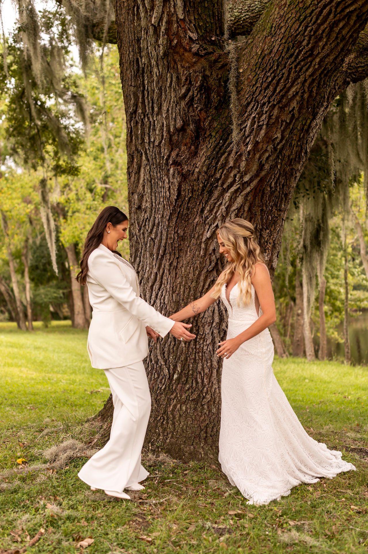 Two people in white dresses playfully dance and twirl around a large tree trunk in a sunny park setting.